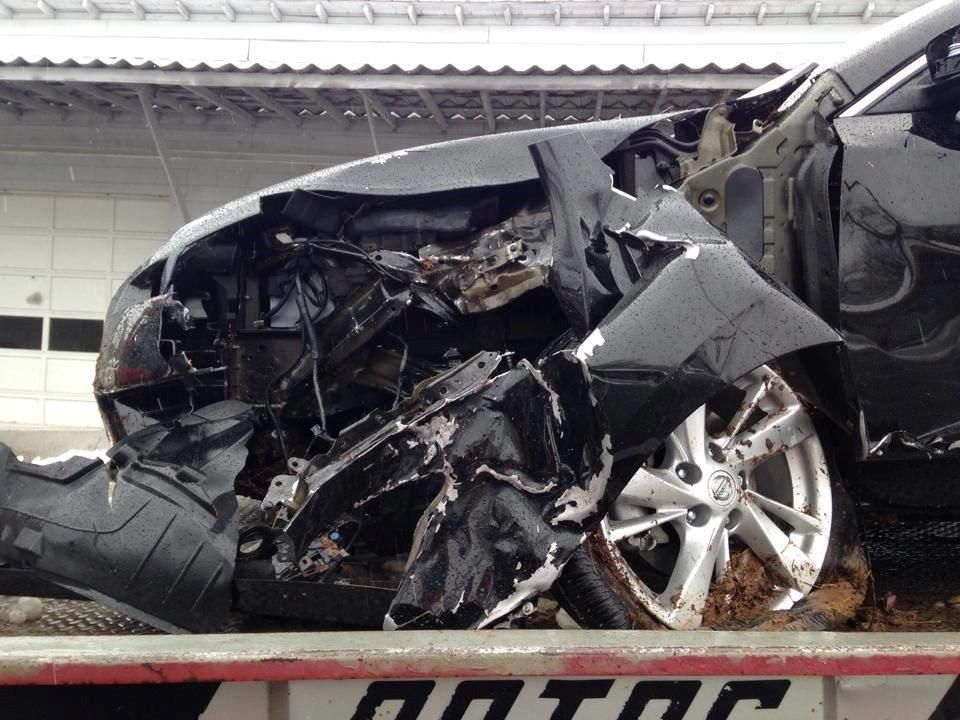 Damaged black car after a collision, front end severely crushed. Tire visible, muddy ground.