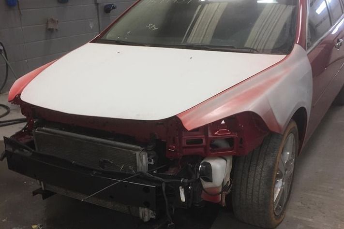 Car undergoing body repair, front-end disassembled, hood and fender primed white, red paint visible.
