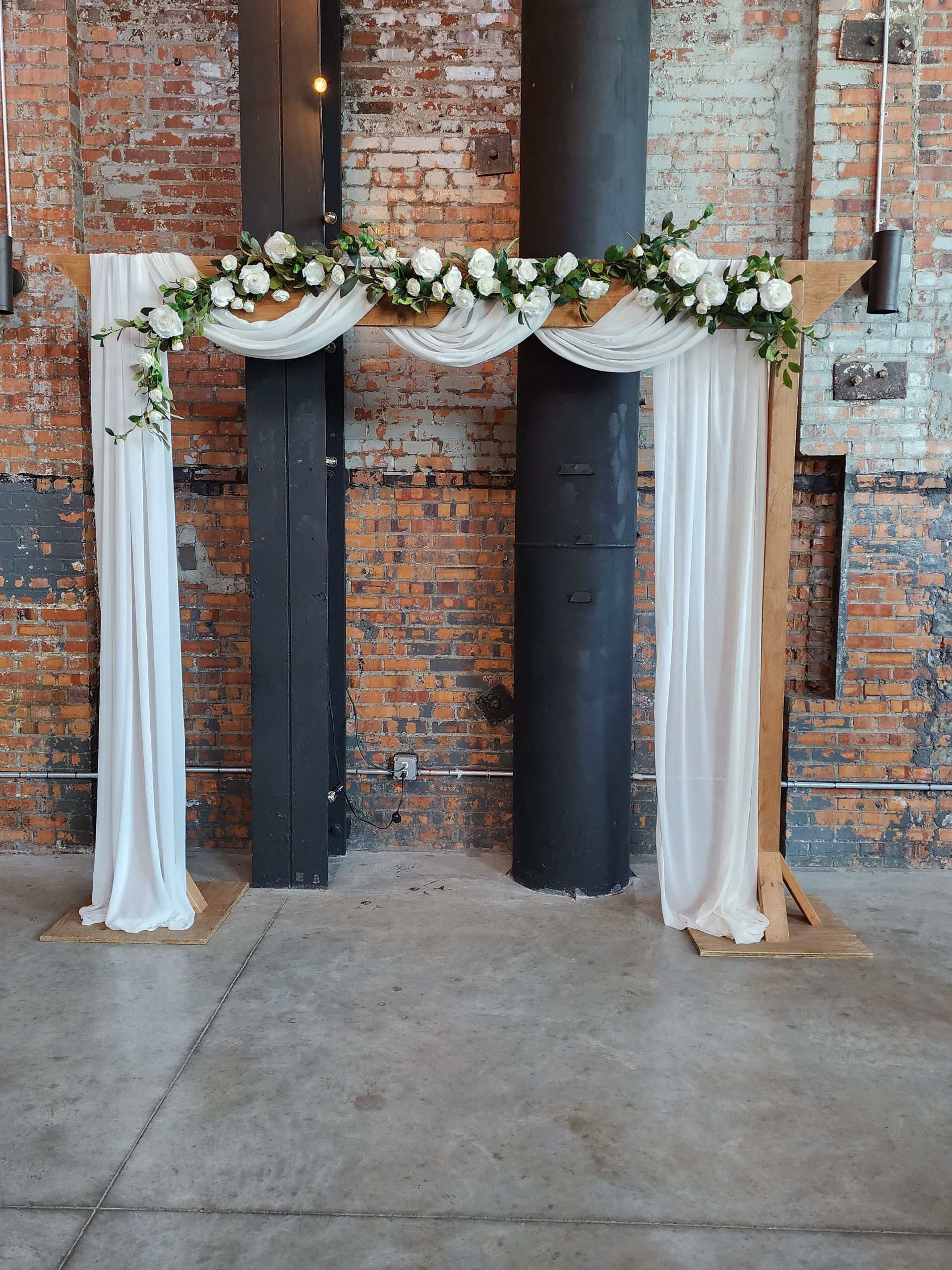 A wooden arch with white curtains and flowers is in front of a brick wall.