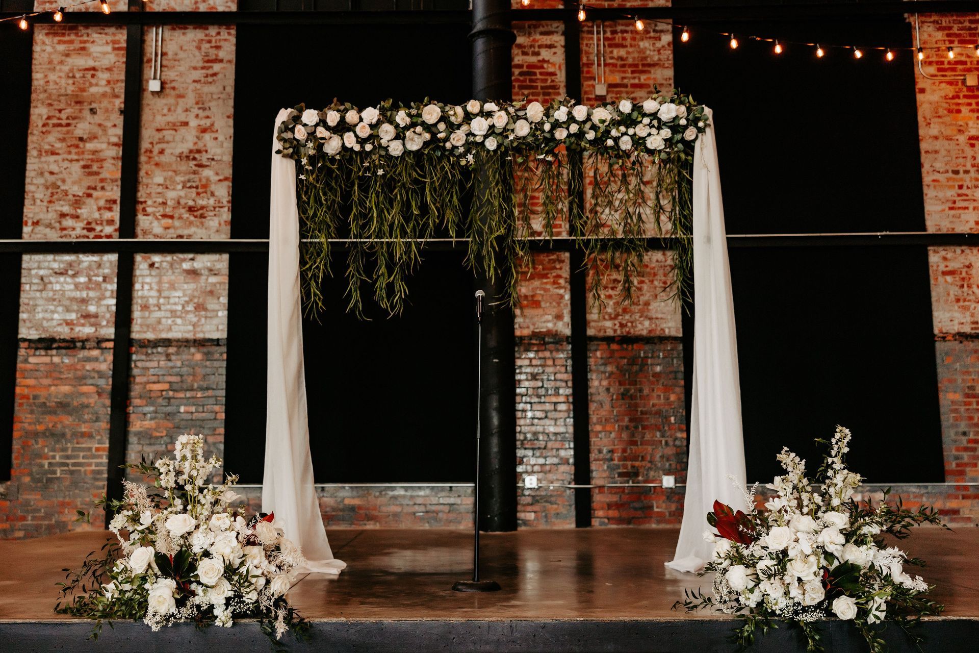 A wedding arch decorated with white flowers and greenery in front of a brick wall.