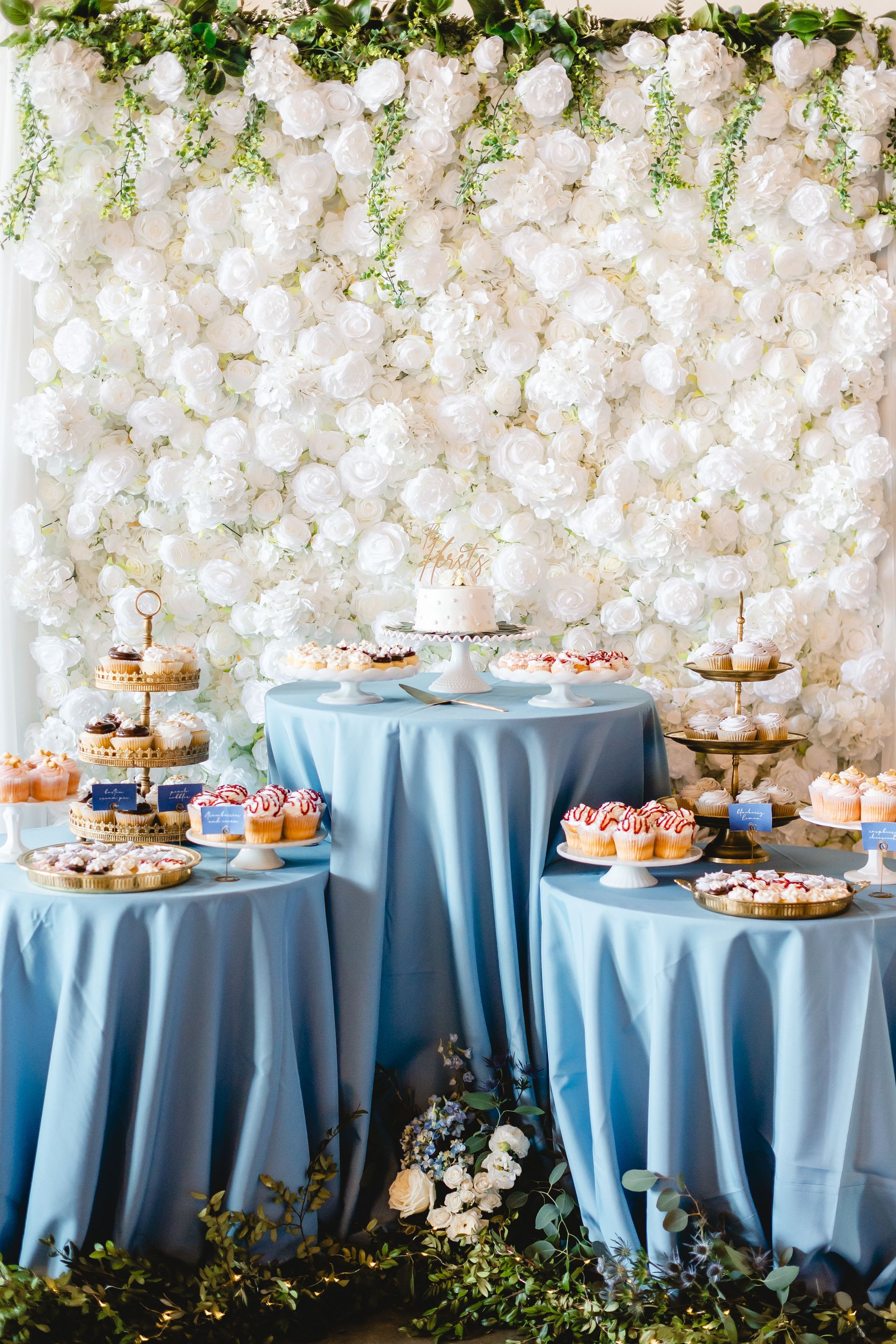 A dessert table with blue tables and a white flower wall behind it.