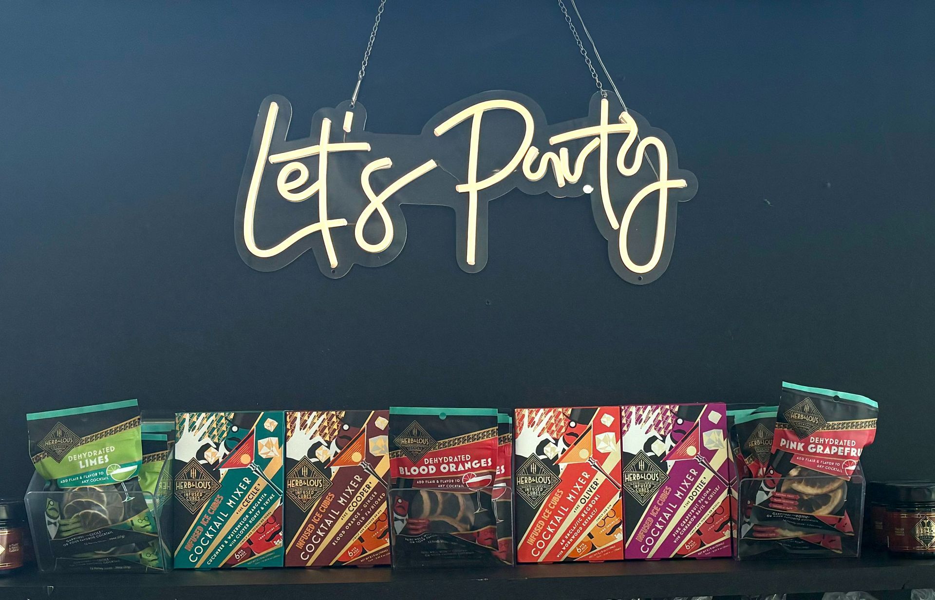 A neon sign that says let 's party hangs above a shelf of candy.