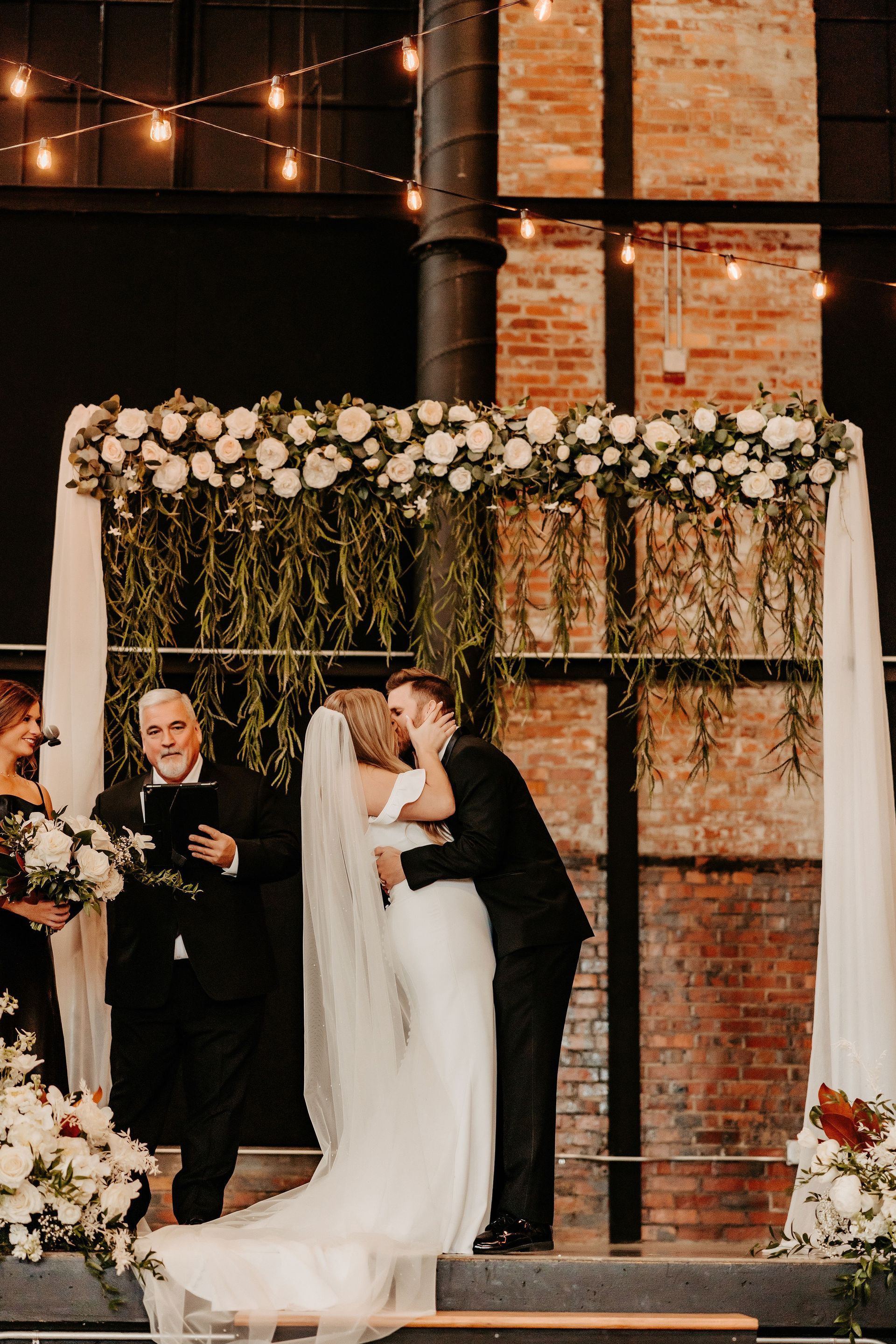 A bride and groom kissing at their wedding ceremony under a floral arch.