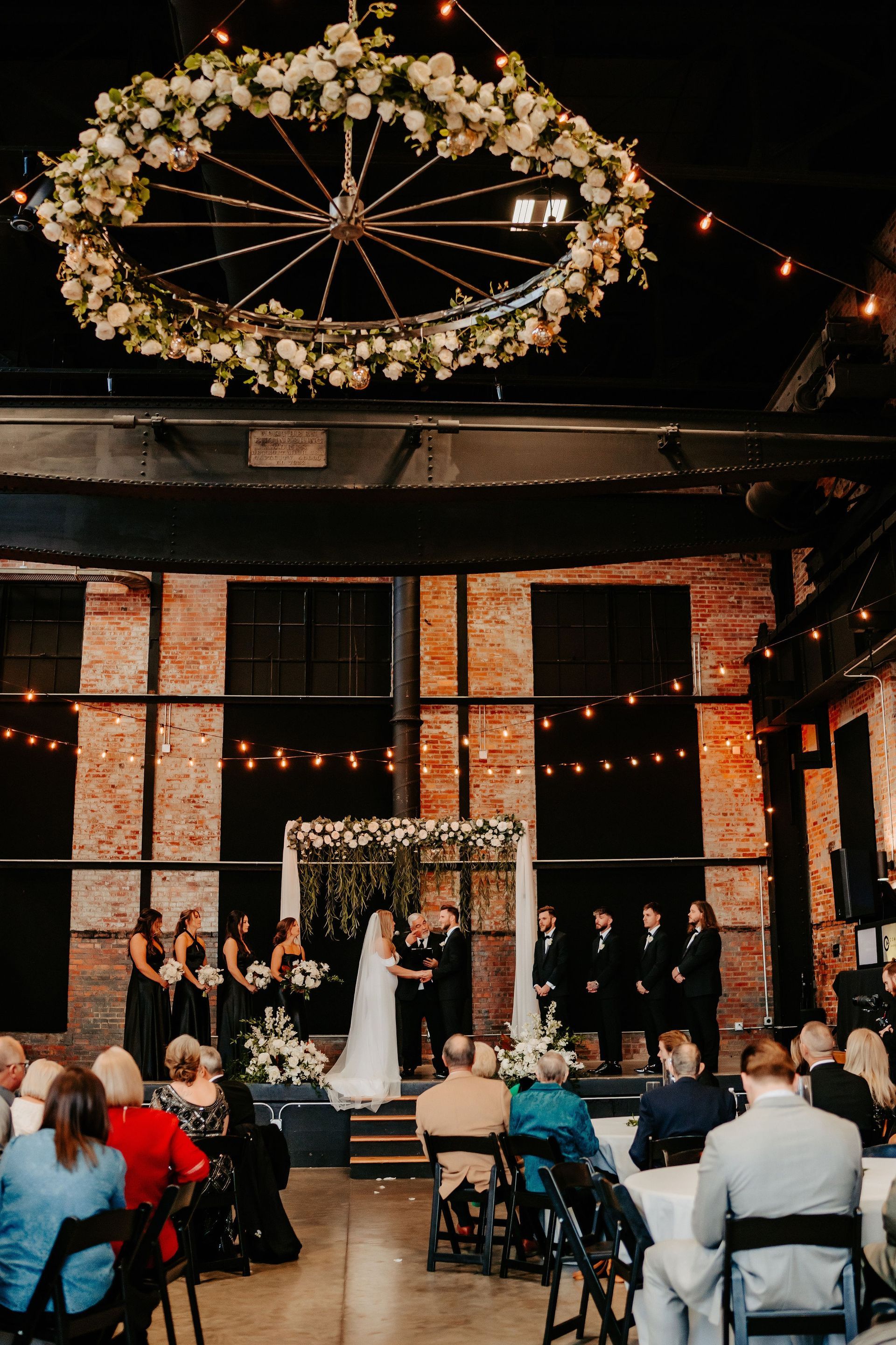 A bride and groom are getting married in a large room with a large wreath hanging from the ceiling.