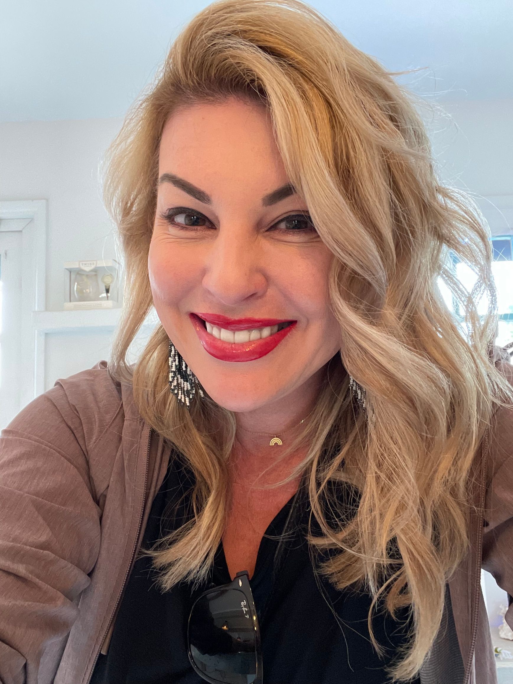 Woman with long, wavy blonde hair smiles widely; wearing a black top and brown sweater, red lipstick.