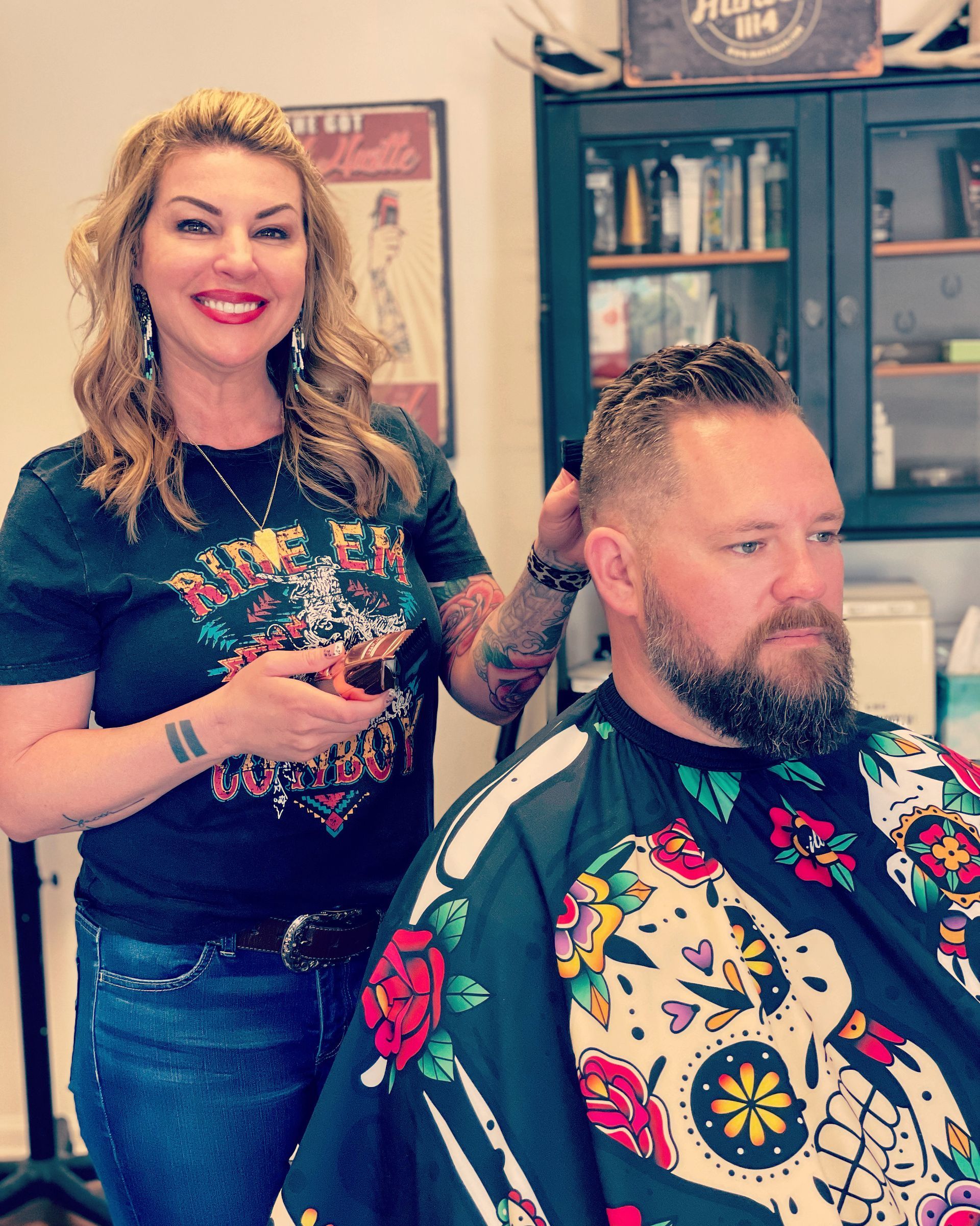 Woman cutting a man's hair in a barber shop. Both are smiling. She wears a graphic tee, he has a patterned cape.