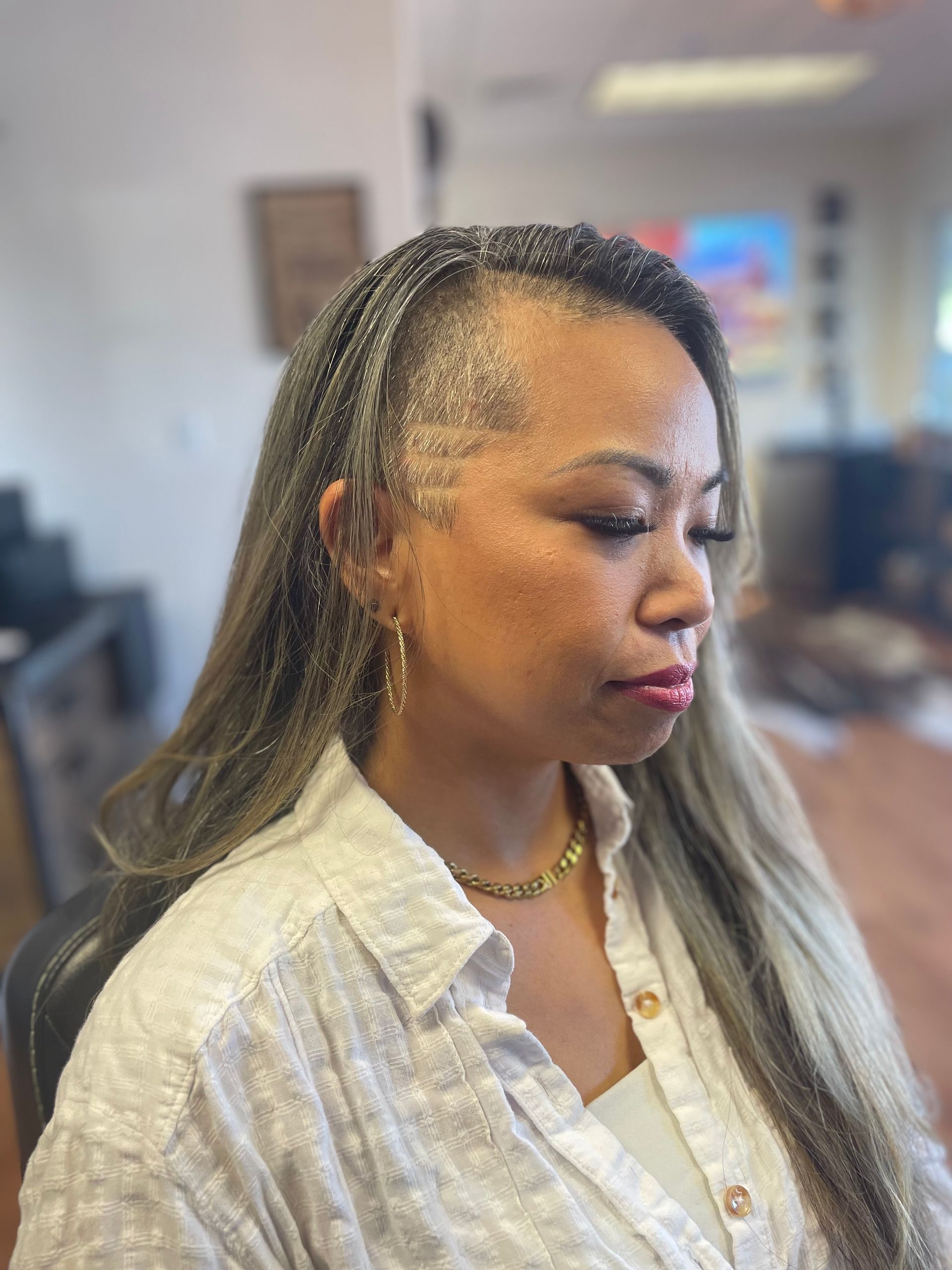 Woman with braided hairline and light hair, wearing a white shirt and gold jewelry, indoors.