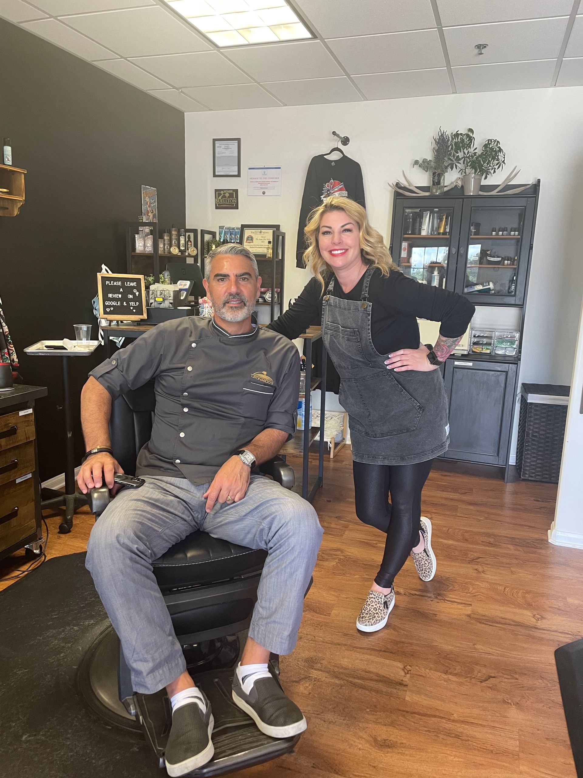 Man seated in barber chair, woman standing next to him, posing inside a barber shop.