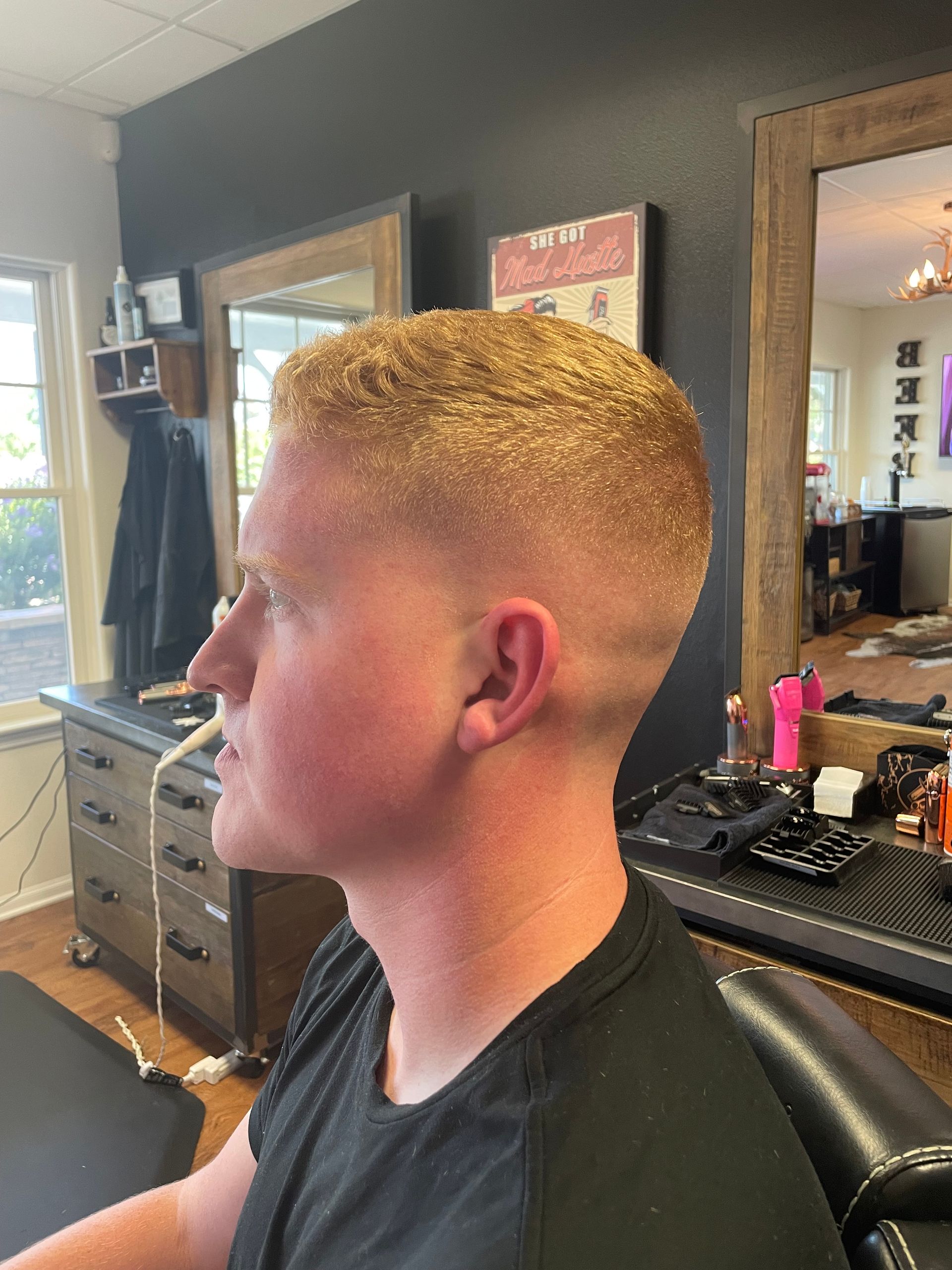 A man with short, faded blonde hair sits in a barber chair. He is in a barbershop.