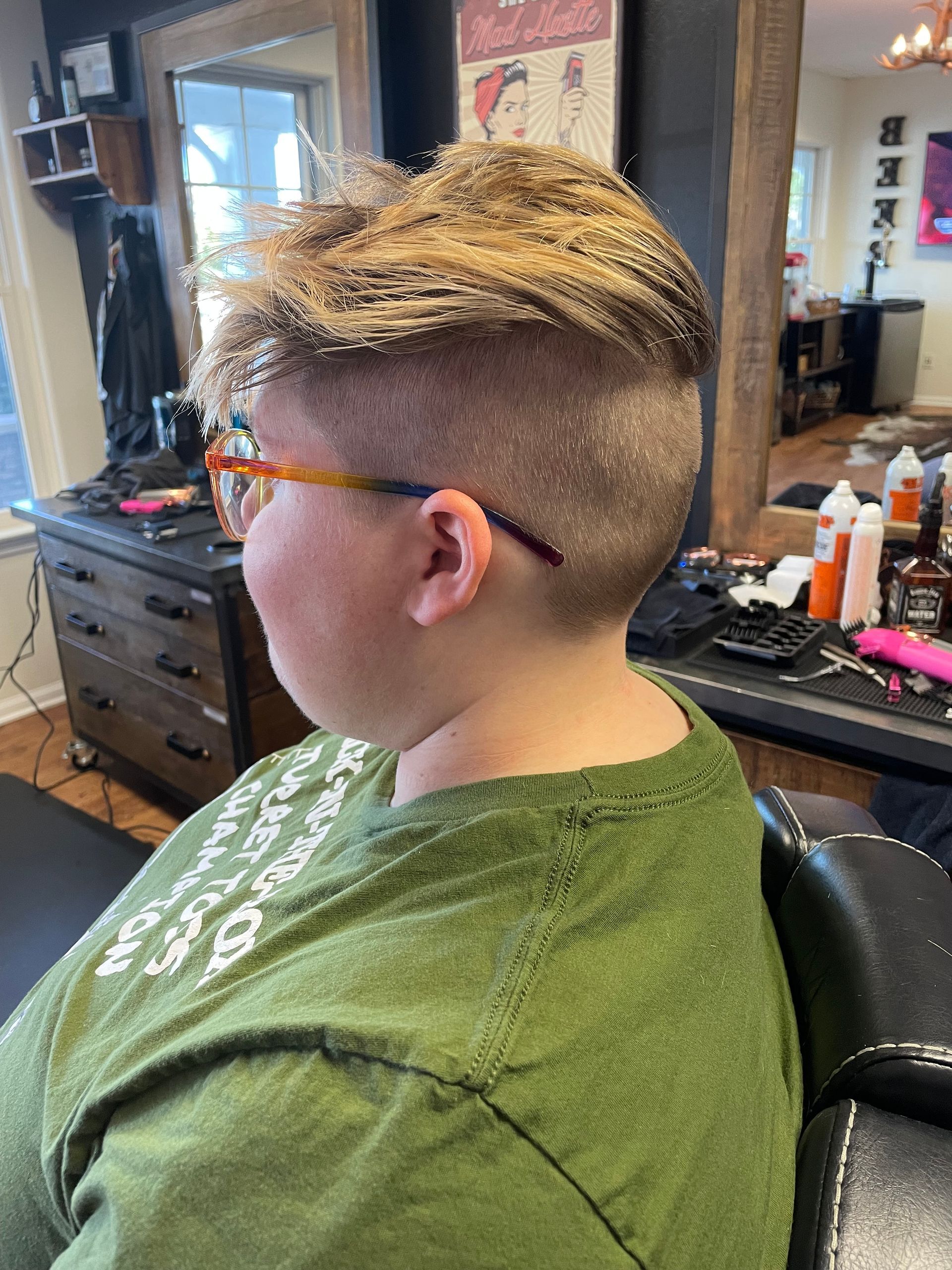 Person with a short haircut and glasses sits in a salon chair, wearing a green shirt.