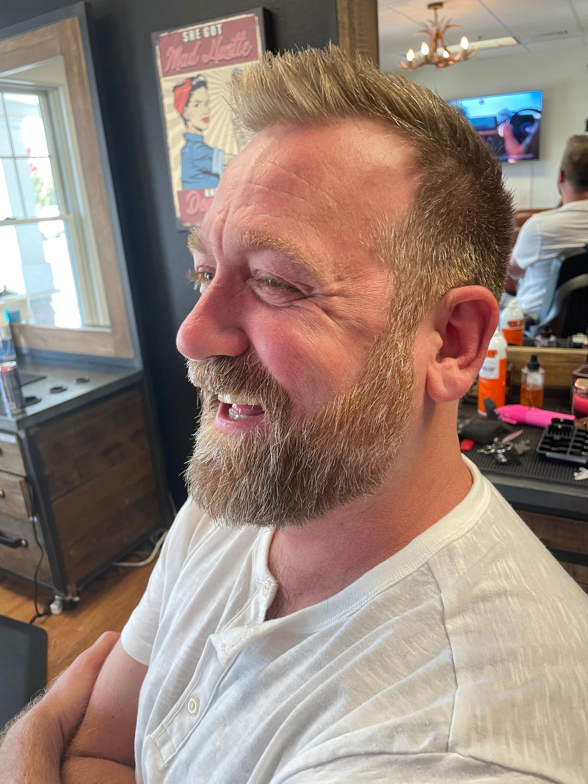 Man with light brown hair and beard in a barber shop smiling, wearing a white shirt.