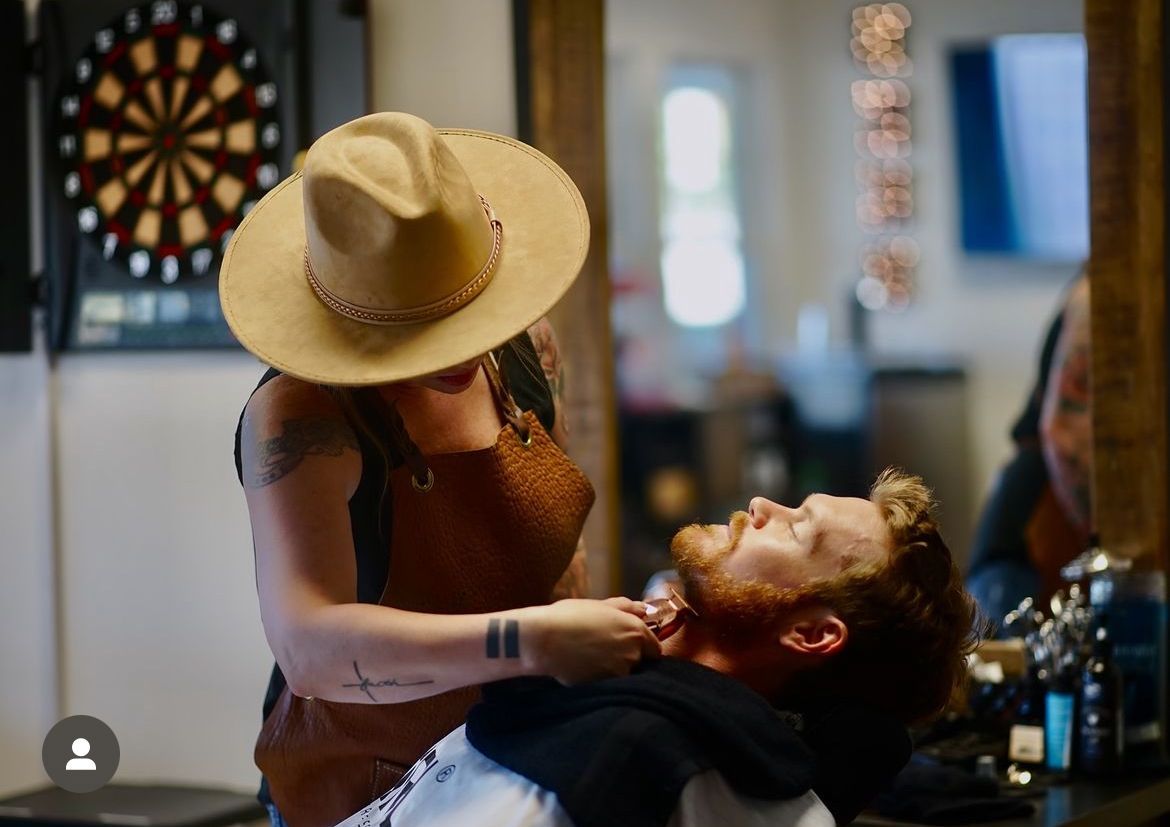 Barber wearing a hat shaves a client's beard in a barbershop.