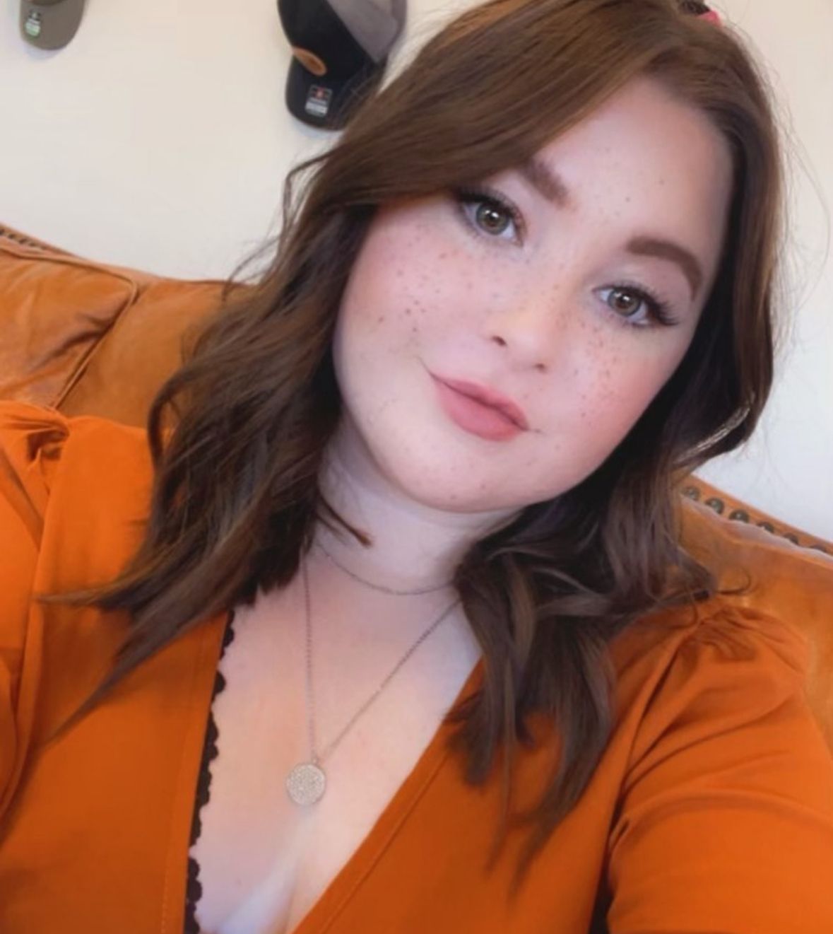 Woman with auburn hair and freckles, wearing an orange top, smiles at the camera on an orange couch.