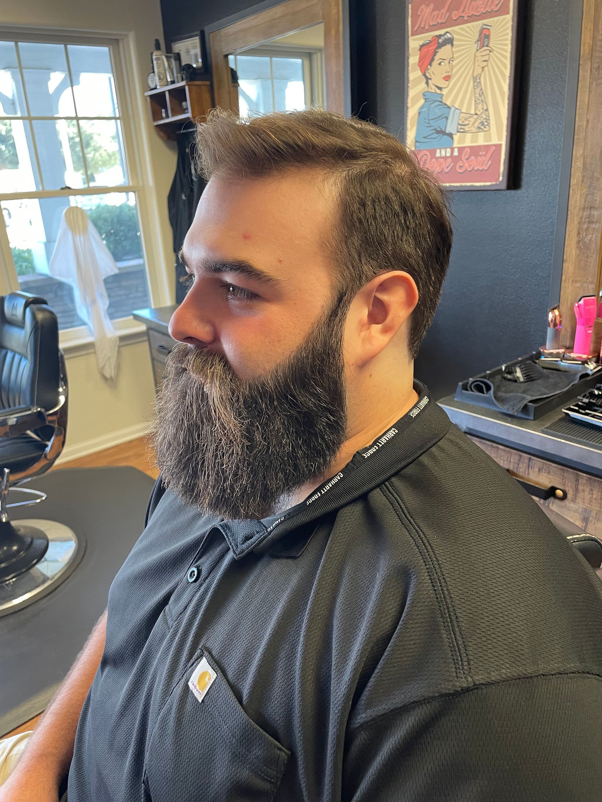 Man with dark beard, in a salon chair, wearing a black smock, looking to his left, indoors.