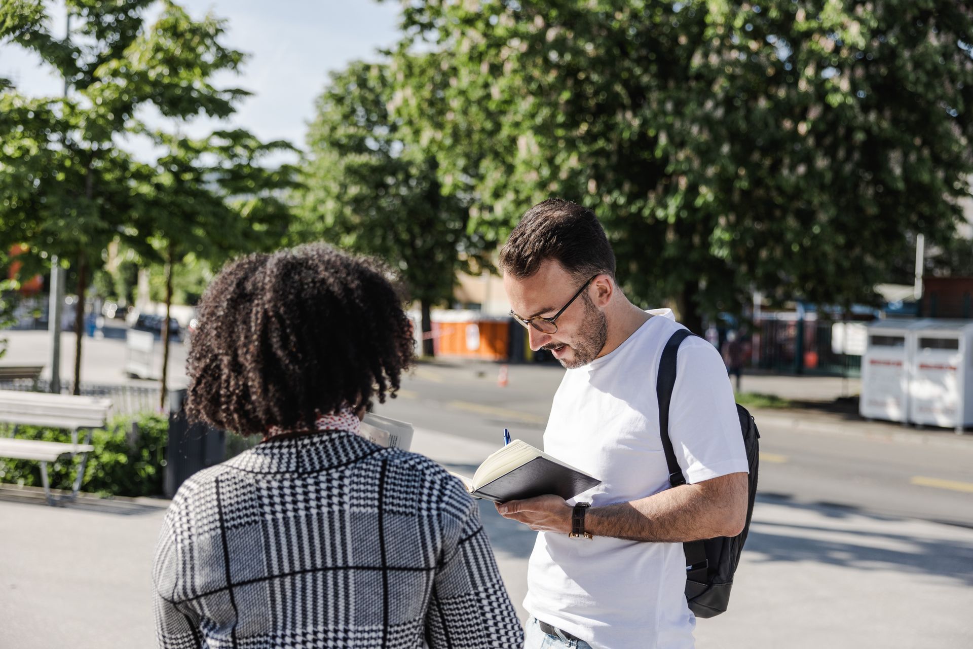Deux personnes se tiennent dehors. L'une porte une veste à carreaux, l'autre des lunettes et un carnet. Des arbres et des bâtiments se dessinent à l'arrière-plan.