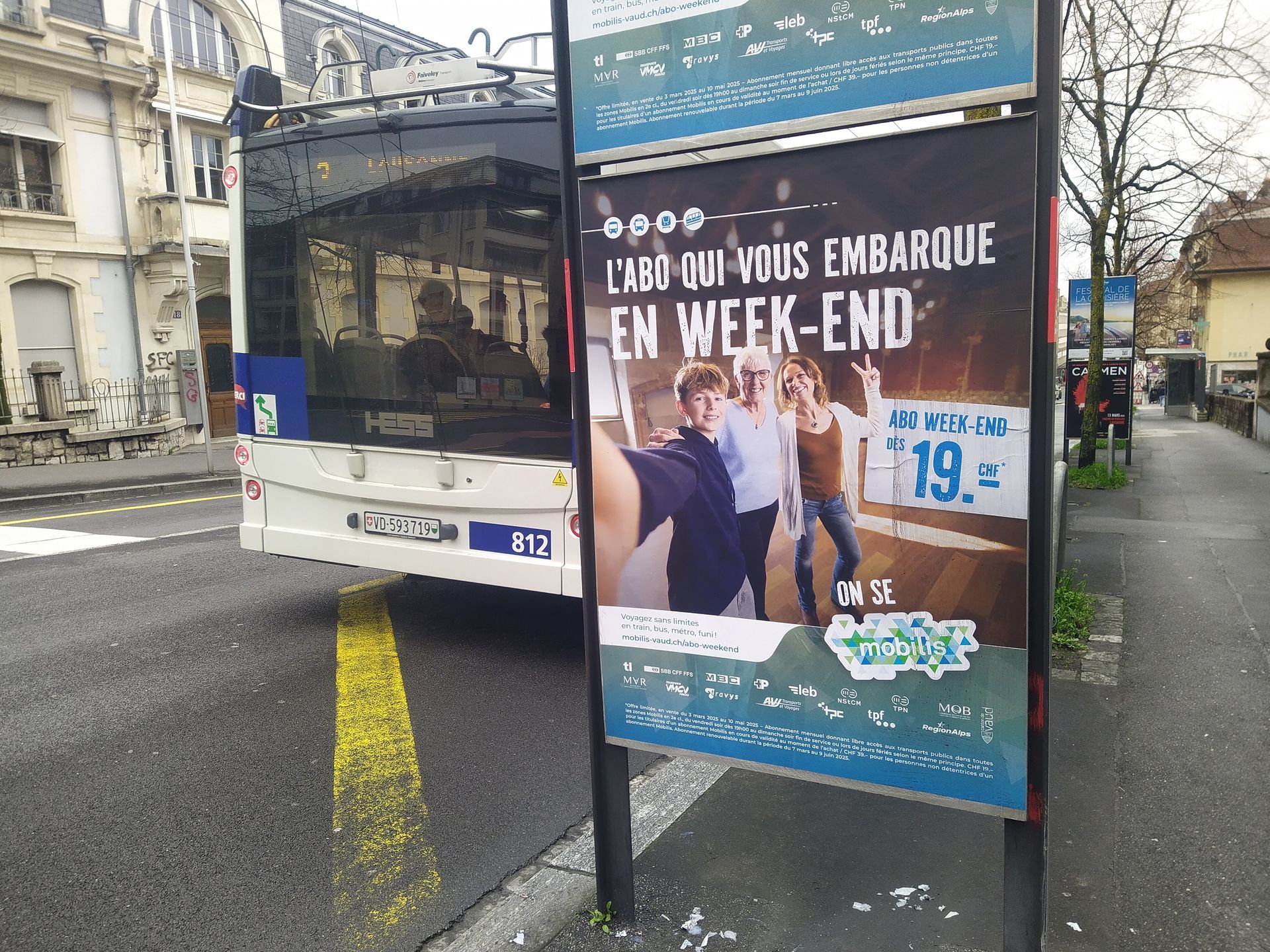 Un bus derrière un kiosque publicitaire bleu et blanc, dans une rue de la ville de Lausanne, avec une affiche promotionnelle pour l'abo weekend de Mobilis