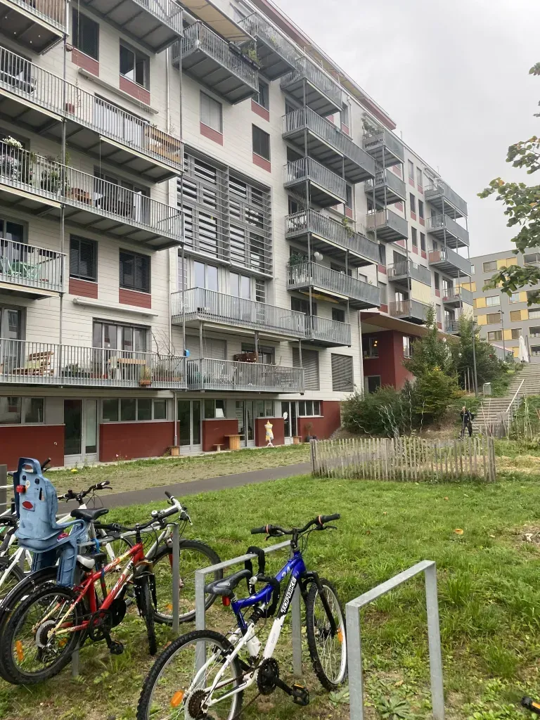Immeuble d'appartements à plusieurs étages avec balcons, vélos garés devant, espace vert et ciel nuageux.