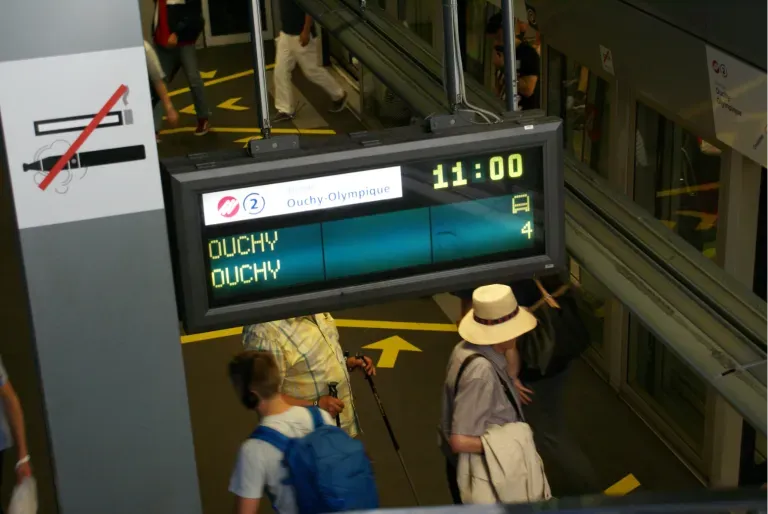 Affichage numérique dans une gare : « 11:00 », destination « OUCHV ». Des personnes passent devant un panneau « Interdit de fumer ».