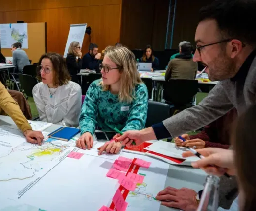 Des personnes assises à une table consultent des cartes et des post-it pendant une réunion. Un homme montre du doigt ; les autres écoutent.