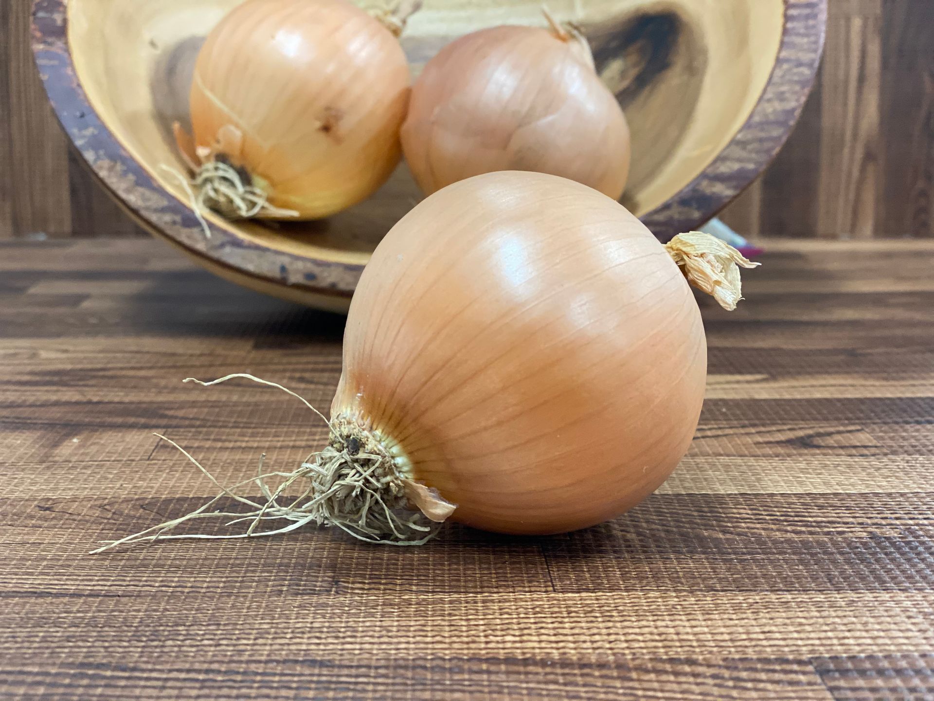 Three yellow onions: one in front, two in a wooden bowl, on a wood-grain surface.