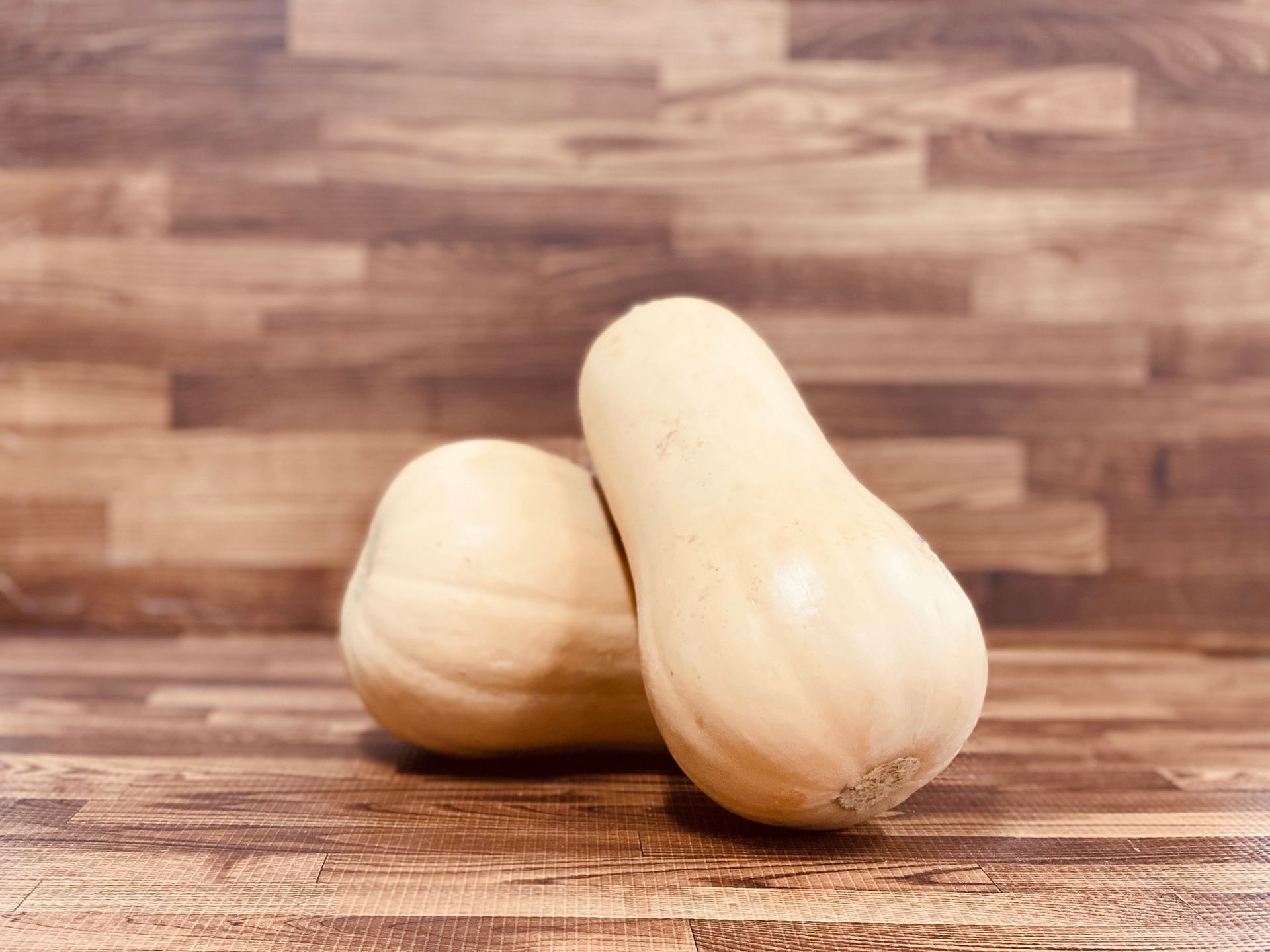 Two butternut squashes on a wood-grain surface. They are a light beige color.