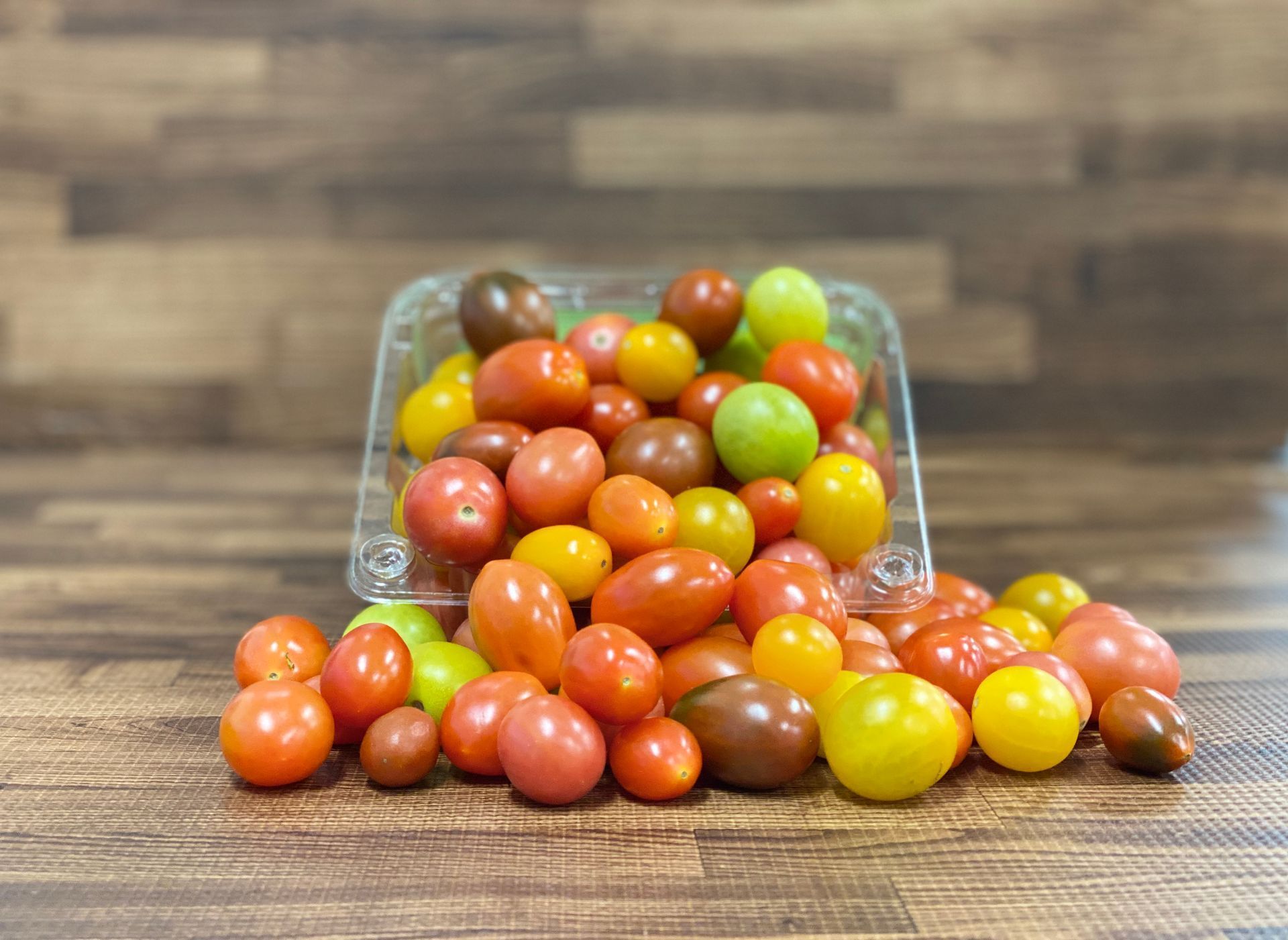 Cherry tomatoes spilling from a clear plastic container on a wood surface. Varied colors: red, yellow, green, and brown.