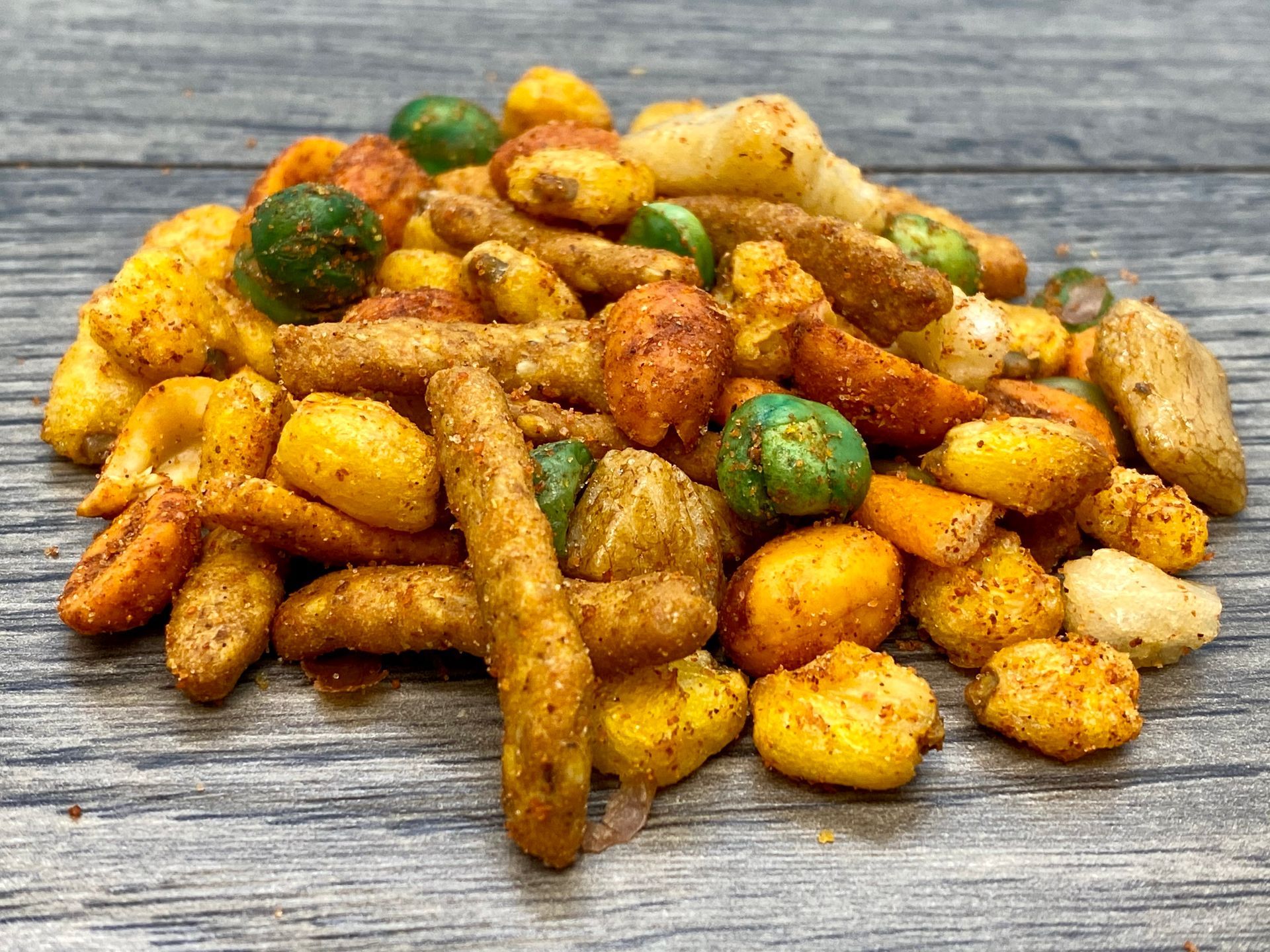 Spicy snack mix on a weathered wooden surface. Contains nuts, corn, and various crunchy shapes, with orange and green accents.