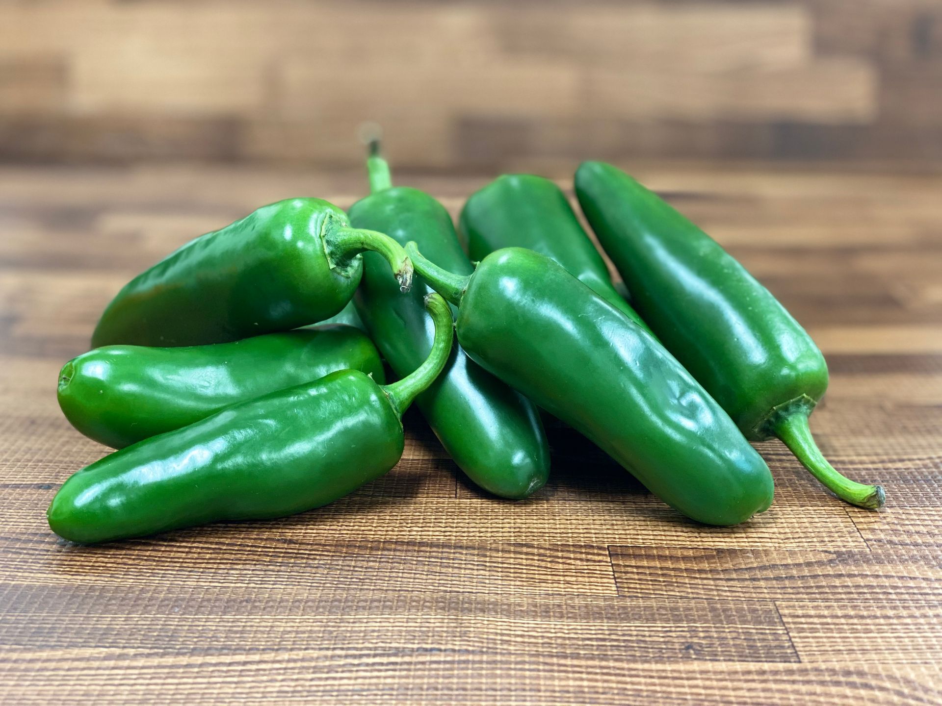 Pile of glossy, bright green jalapeno peppers on a wooden surface.