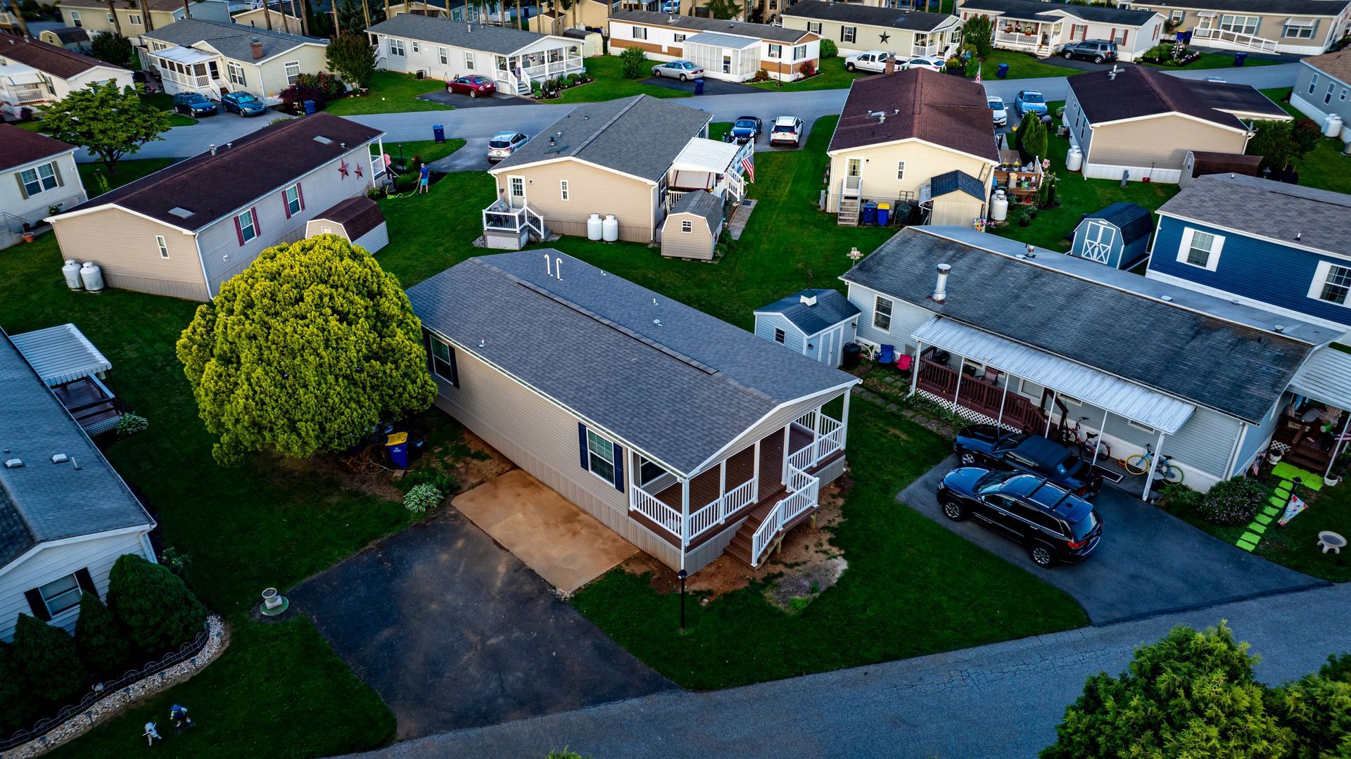 Aerial view of a suburban neighborhood with houses, driveways, parked cars, and green lawns at dusk