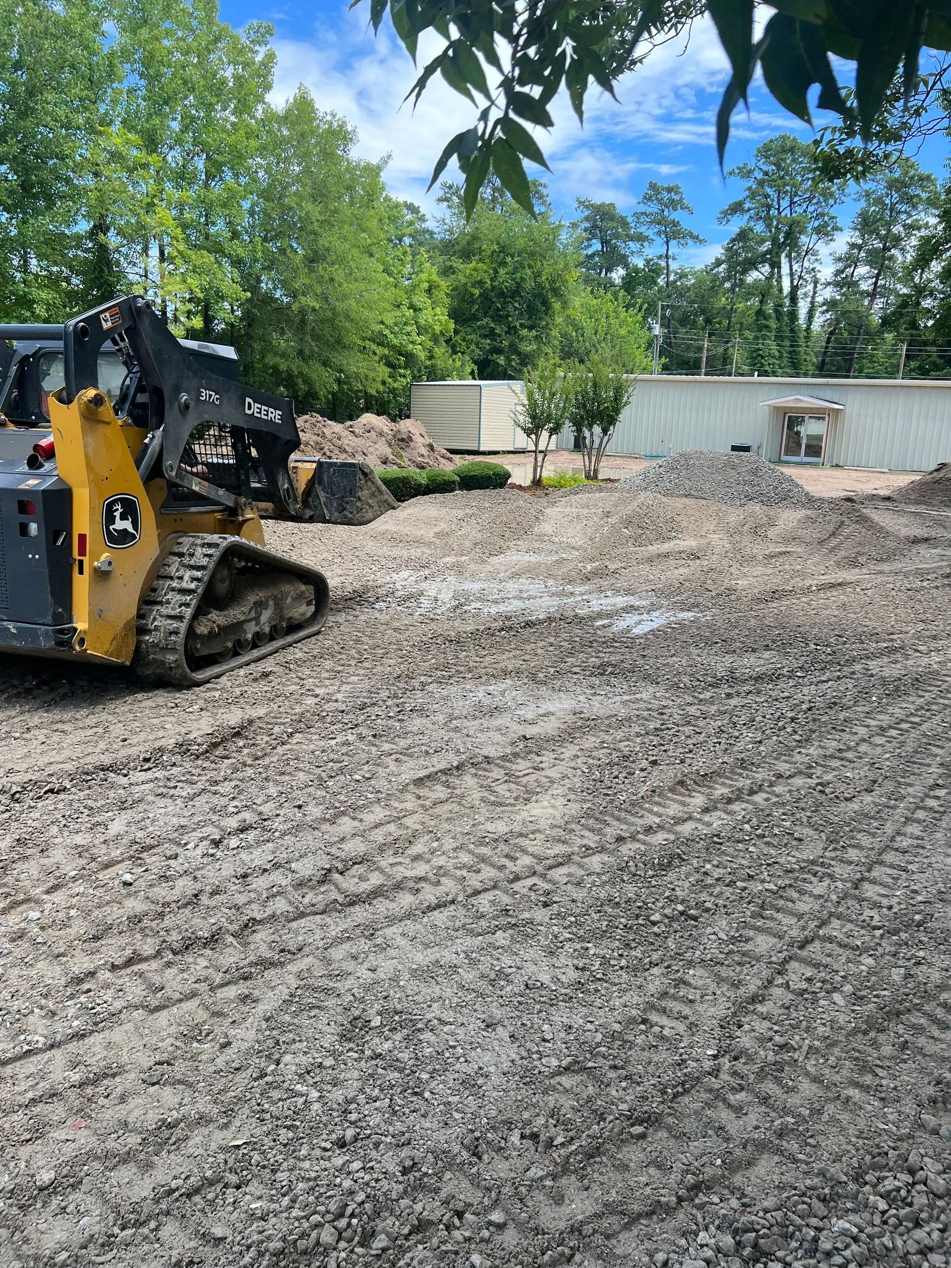 A John Deere skid steer on muddy ground. Construction site with dirt piles, trees, and a building.
