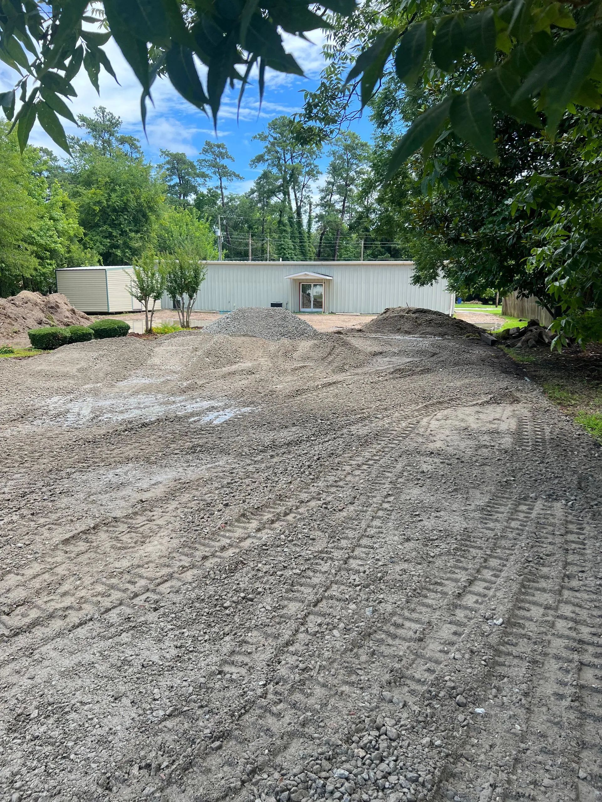 A gravel construction site with tracks leading toward a white rectangular building under a partially cloudy blue sky.