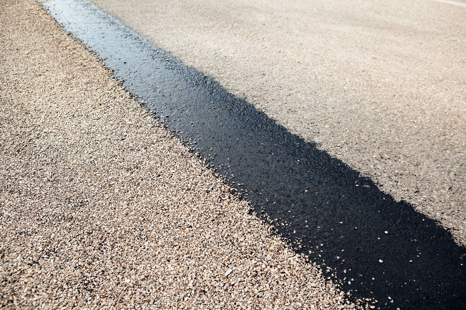 A diagonal view of a road surface showing a dark, freshly applied strip of asphalt bordering a light gravel shoulder.