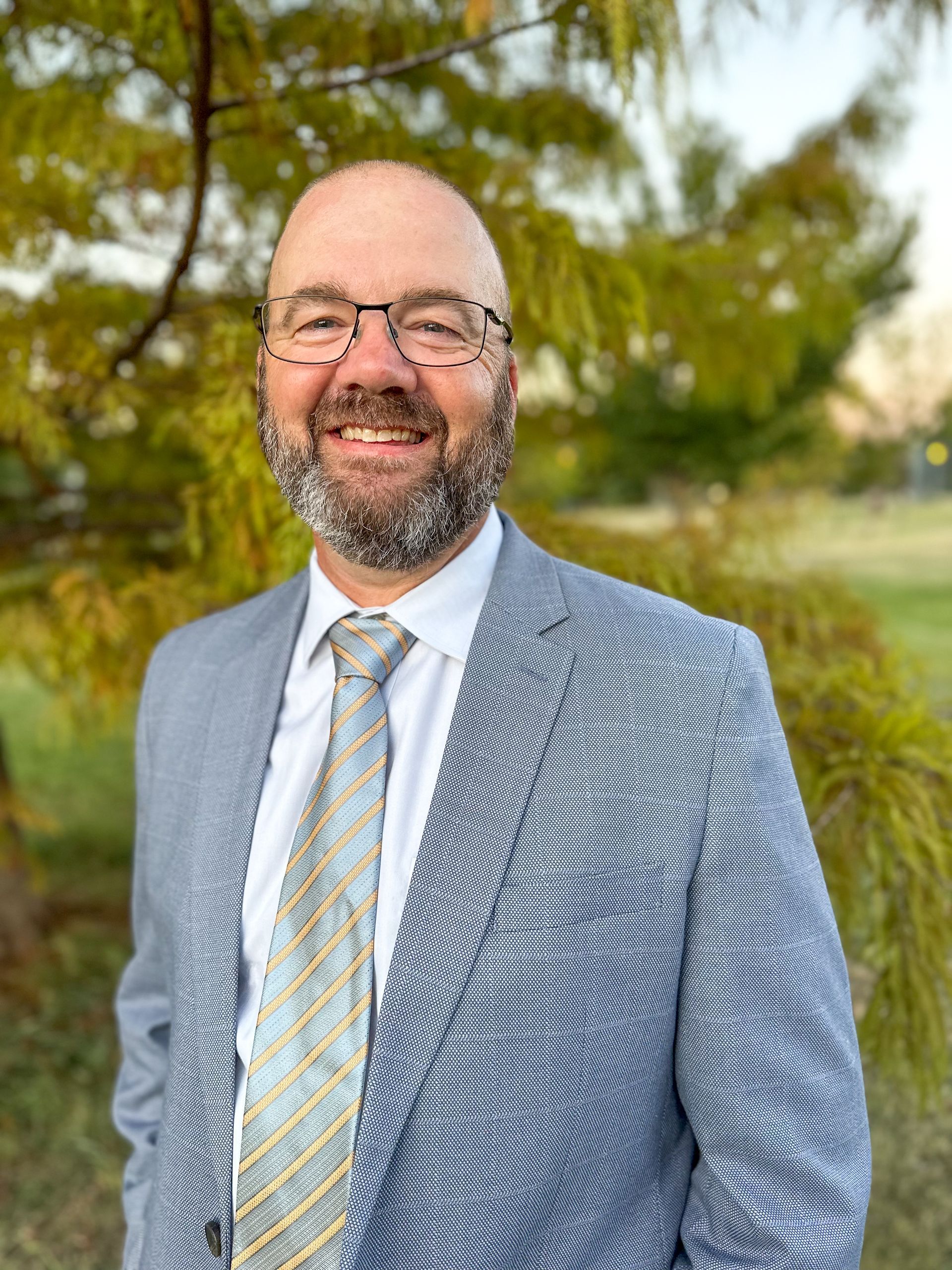 A man in a suit and tie is smiling in front of a tree.