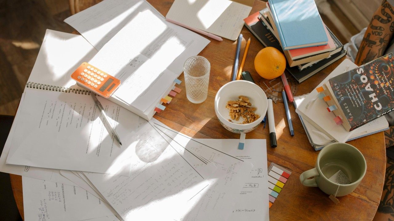 Desk with scattered papers, books, pencils, cup of nuts, and an orange in sunlight.