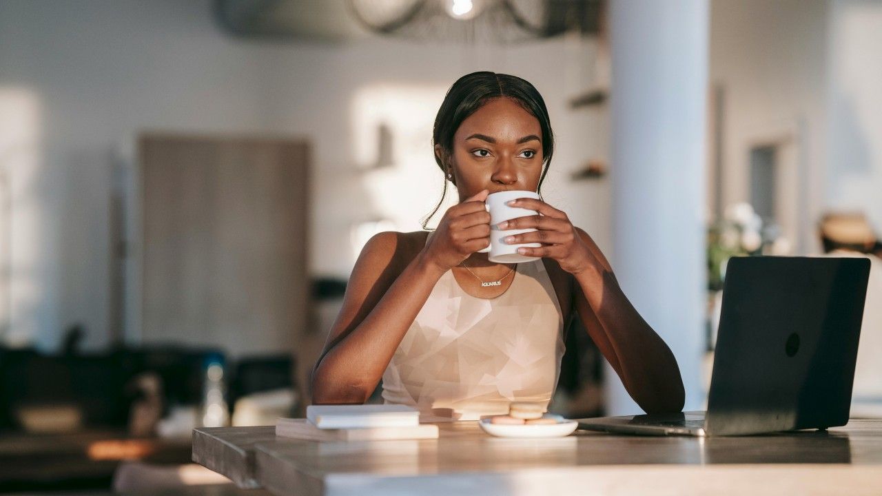 Woman sipping from a mug, working on a laptop at a counter with sunlight.