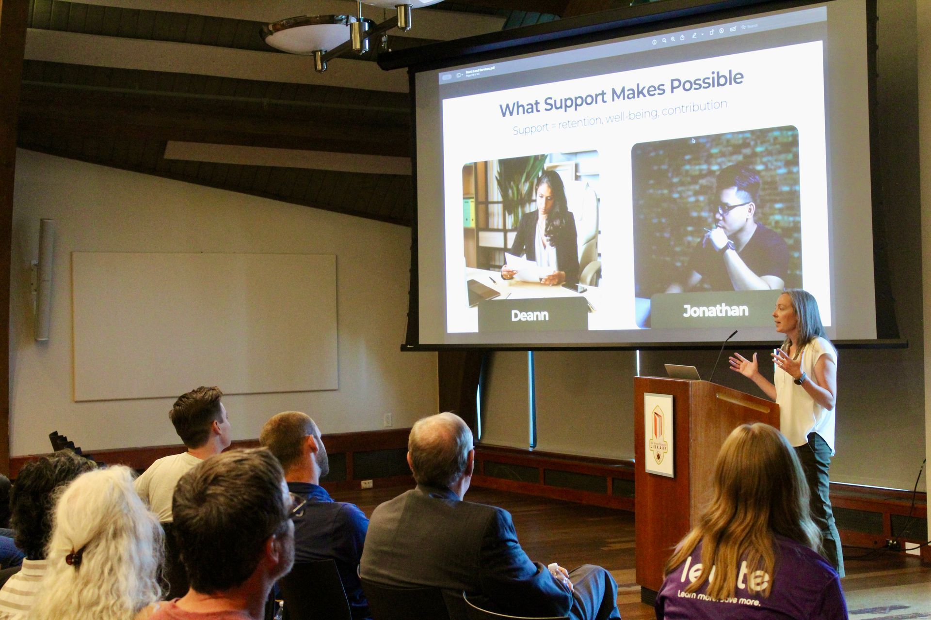 A woman speaks at a lectern in a presentation, with a screen showing two headshots. Audience members sit and watch.