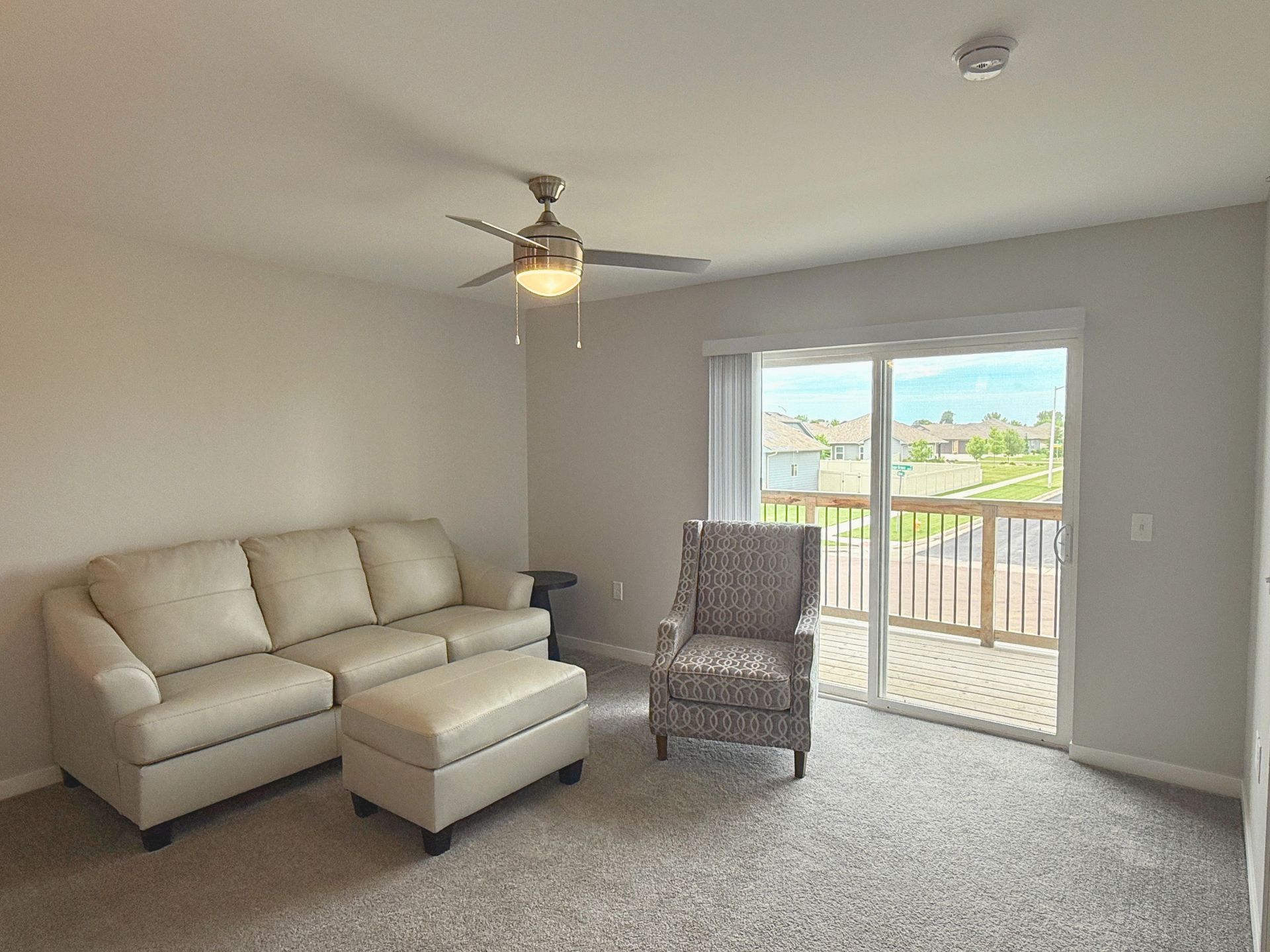 Bright living room with beige sectional, wicker chair, ceiling fan, and balcony door to a sunny view