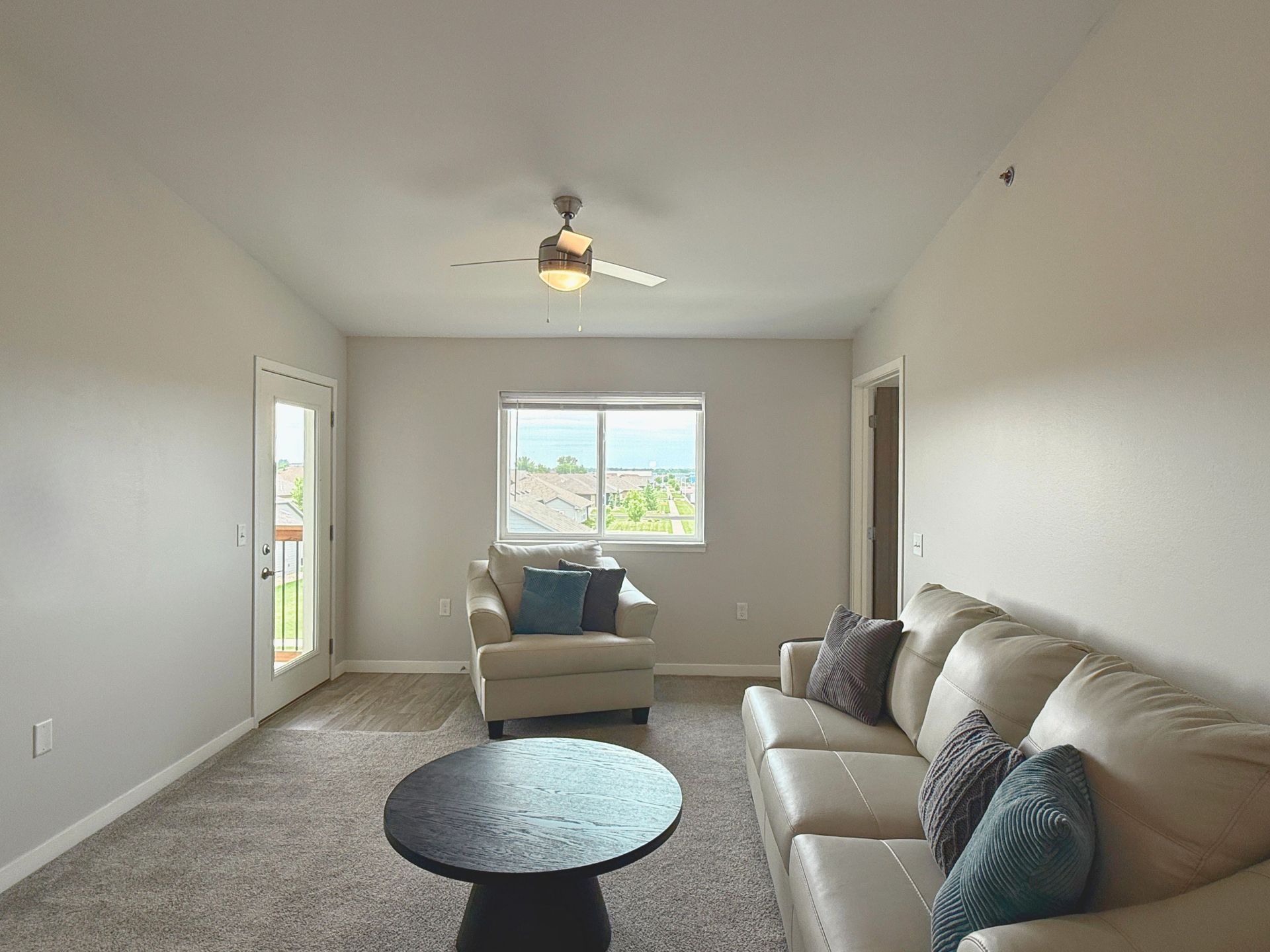 Empty beige living room with couch, ottoman, round coffee table, and large window.