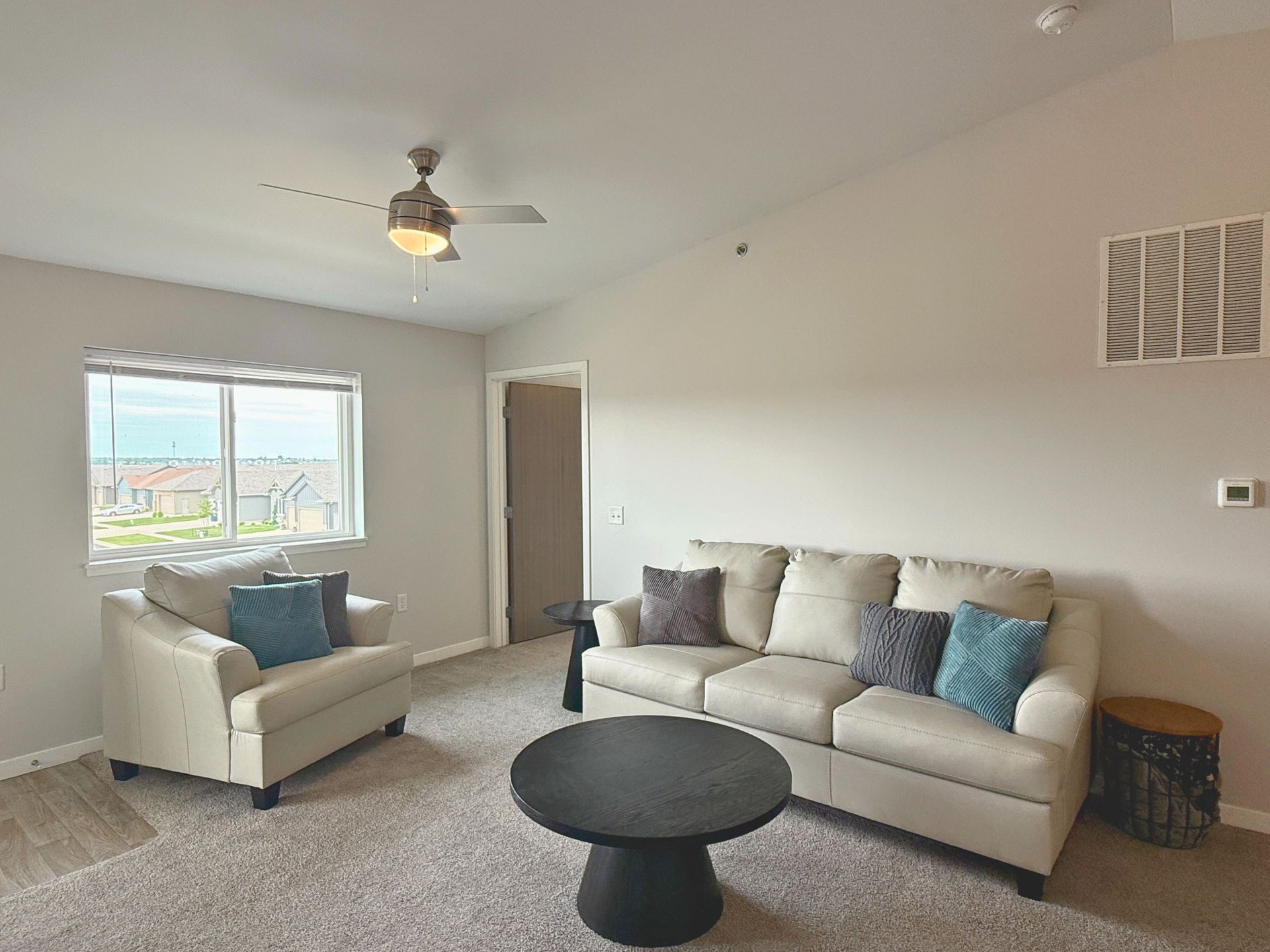 Bright living room with beige sofa and armchair, black coffee table, window, and ceiling fan.