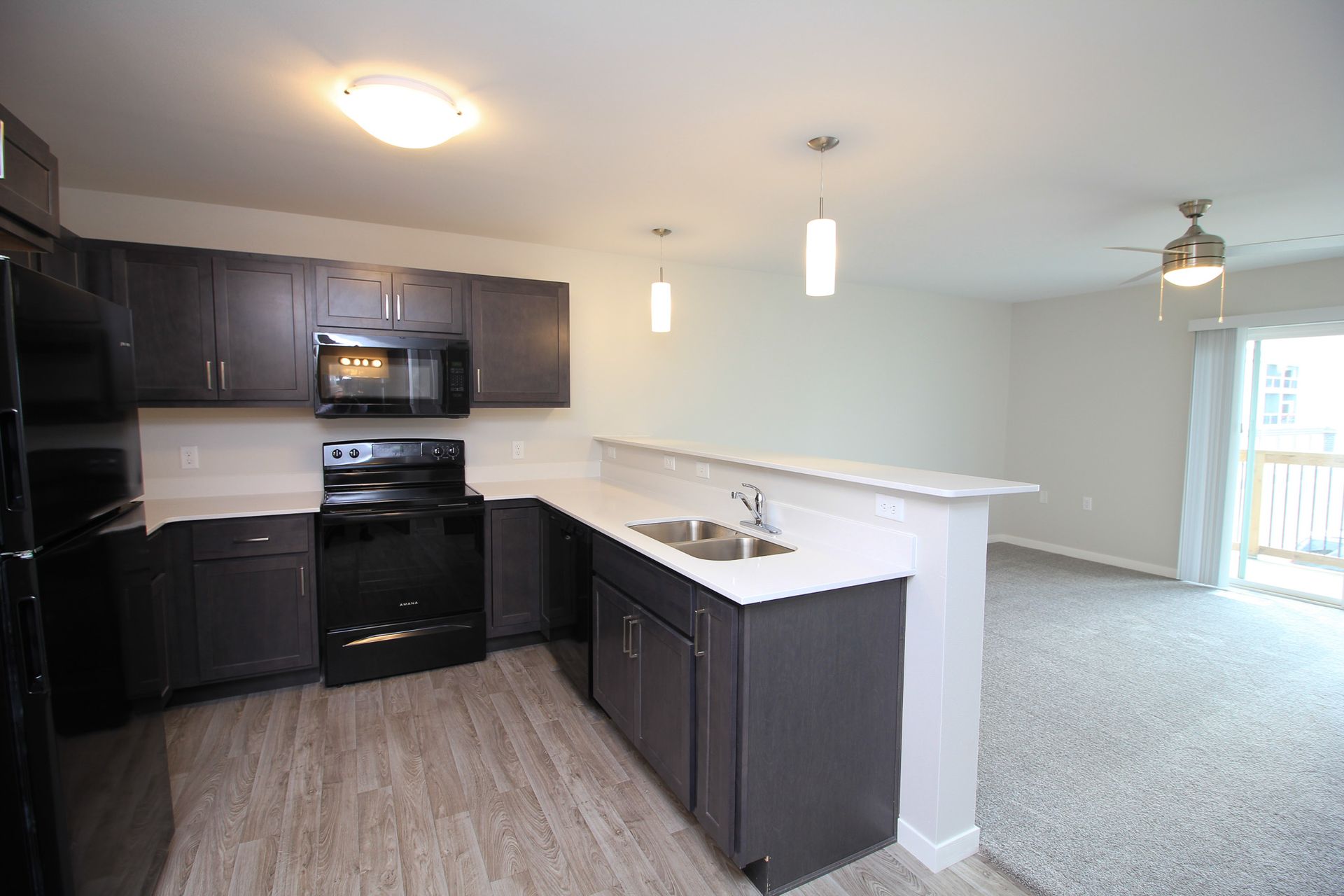 Open-plan kitchen with dark cabinets, black appliances, white breakfast bar, and carpeted living area.