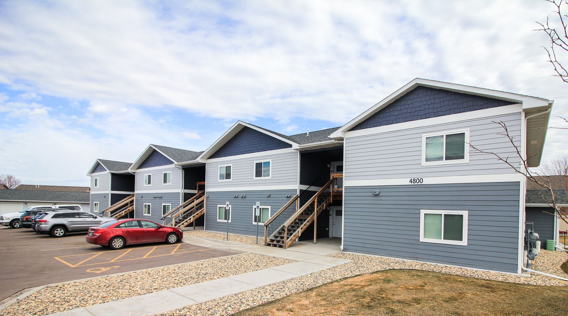 Apartment buildings with blue-gray siding and parked cars along a gravel lot under a cloudy sky
