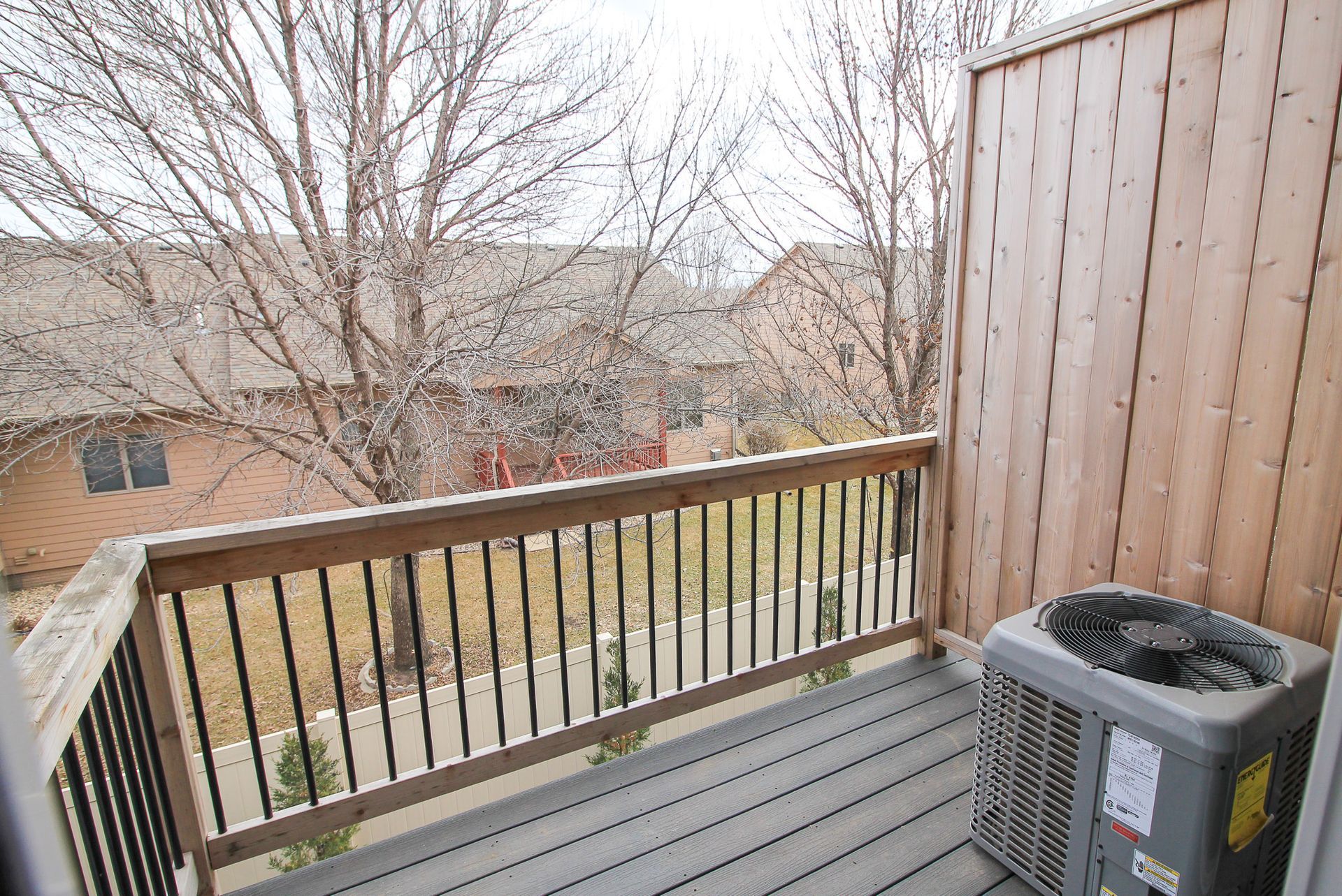 Balcony with wooden railing, gray floor, and an outdoor AC unit overlooking leafless trees and houses.