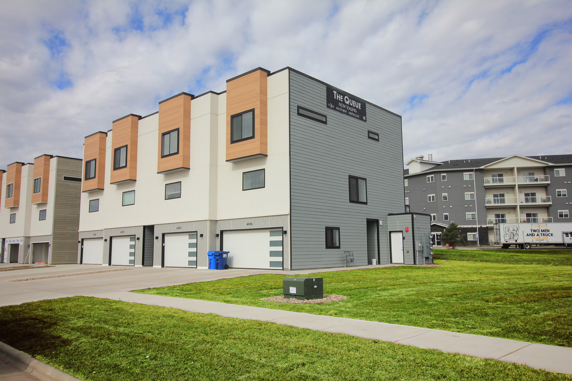 Modern townhouse row with garages, gray and peach exterior, beside green lawn under cloudy sky