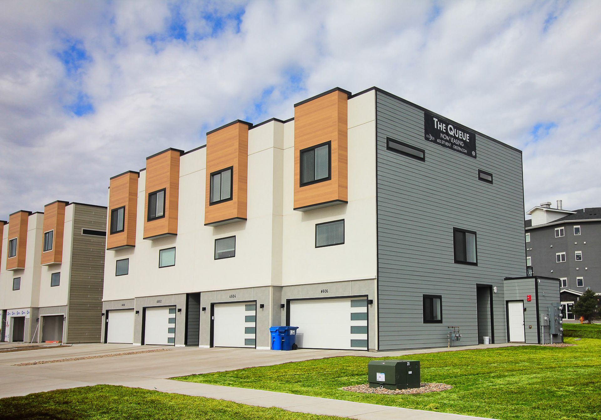 Row of modern townhouses with white and gray walls, orange accents, and a grassy foreground under a cloudy sky