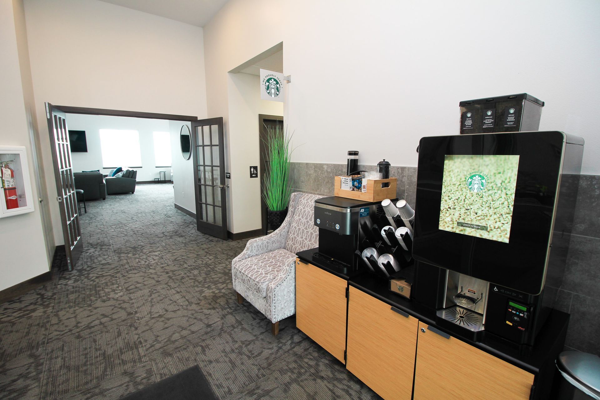 Office lounge with gray carpet, black cabinet, beige seating, and a doorway to a bright conference room