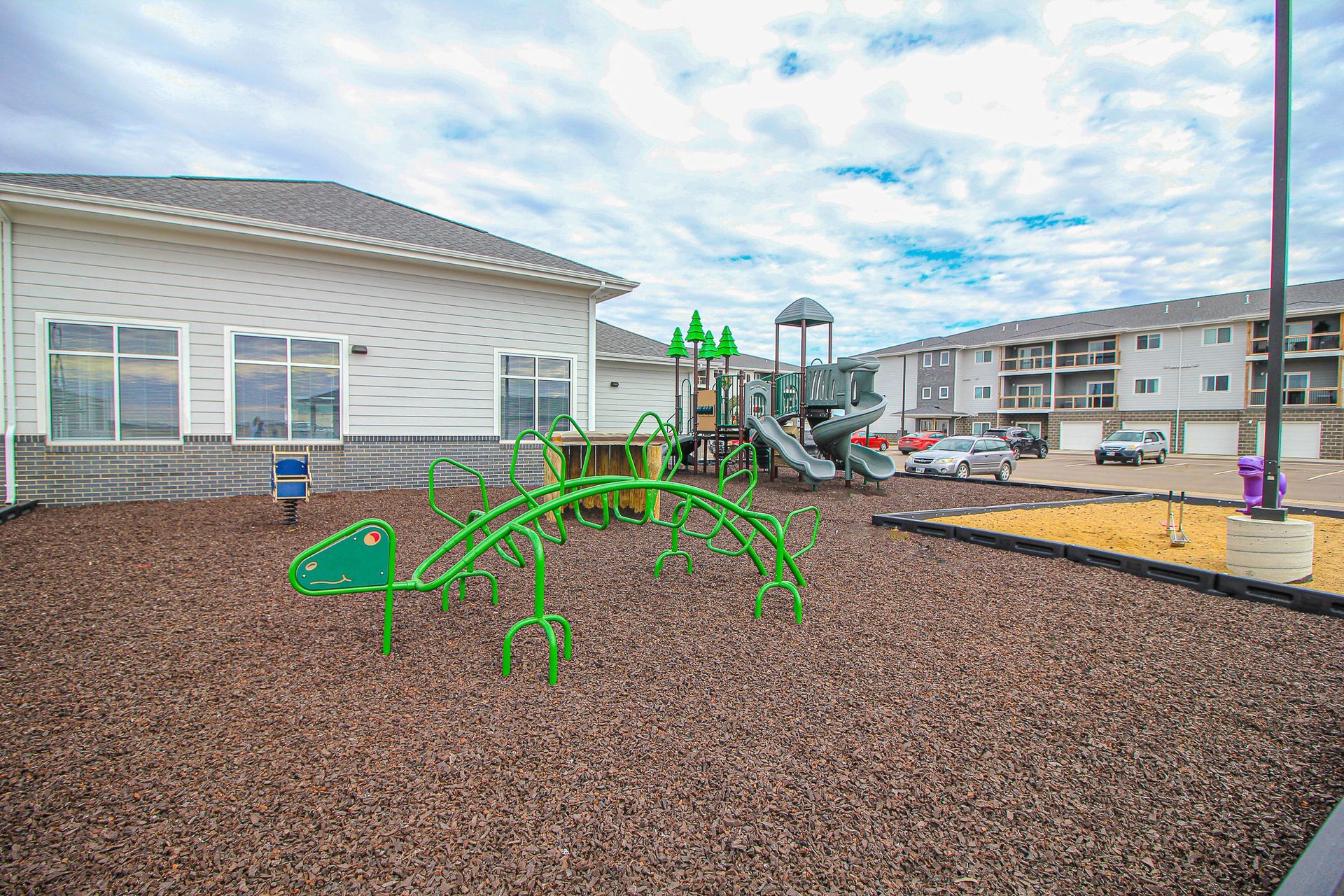 Bright green playground equipment in a mulched courtyard beside apartment buildings under a cloudy sky