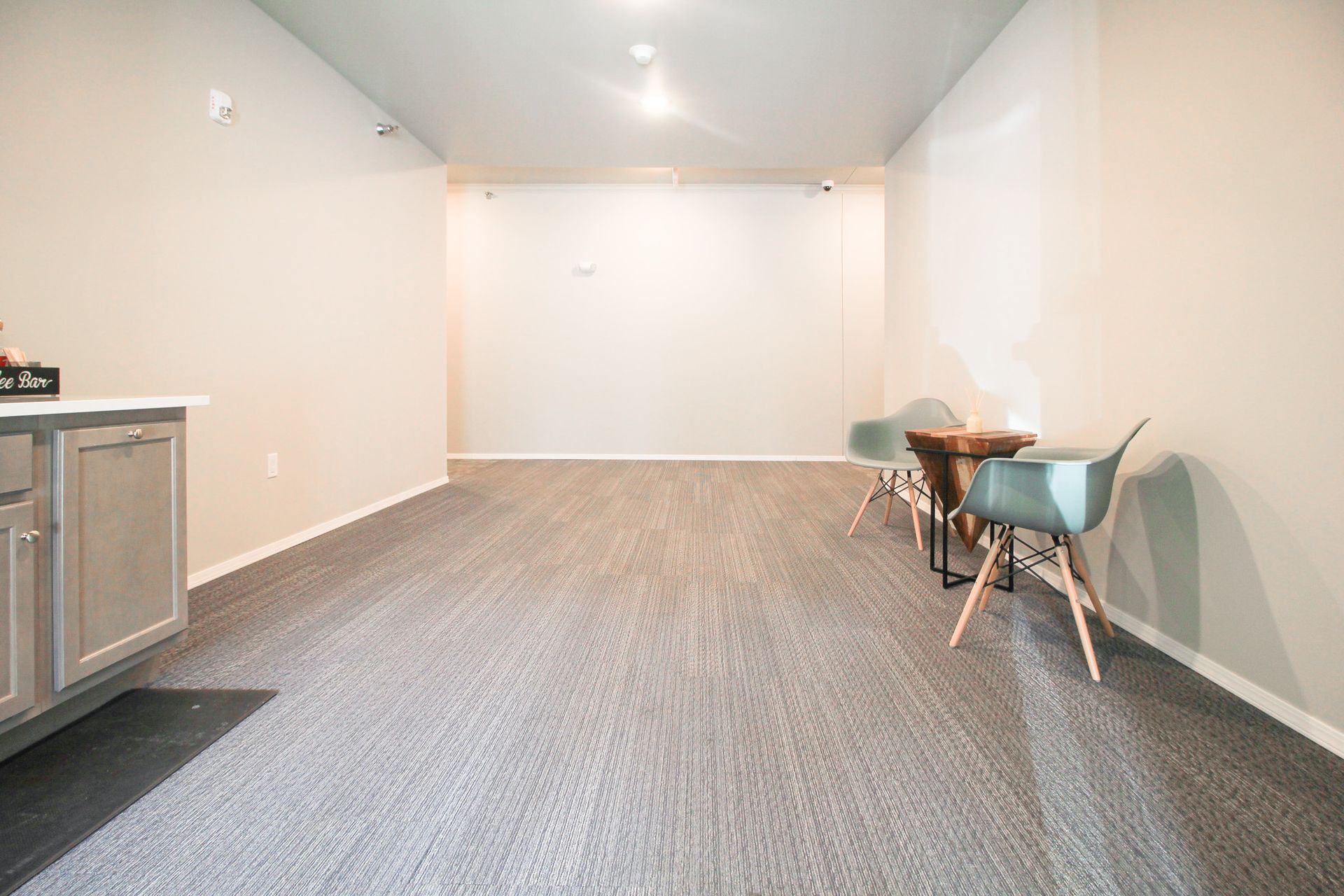 Empty carpeted room with gray walls, a small table and chairs on the right, and a cabinet on the left.