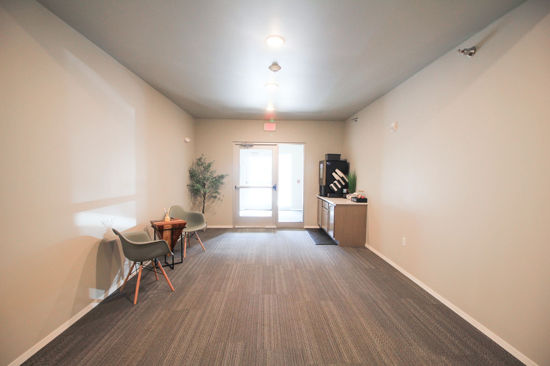 Empty carpeted room with beige walls, a small table and chair, potted plant, and bright French doors at the far end