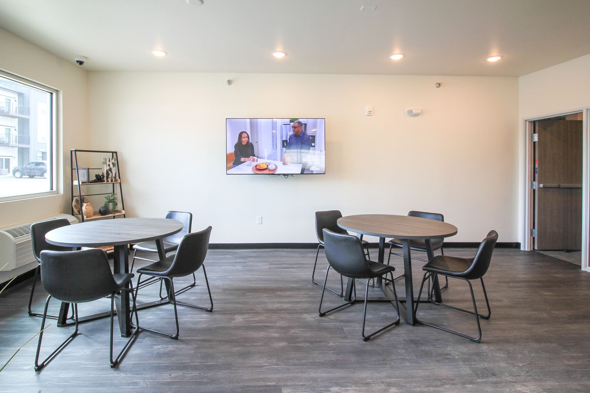 Bright lounge with round tables, black chairs, wall-mounted TV, and large windows.