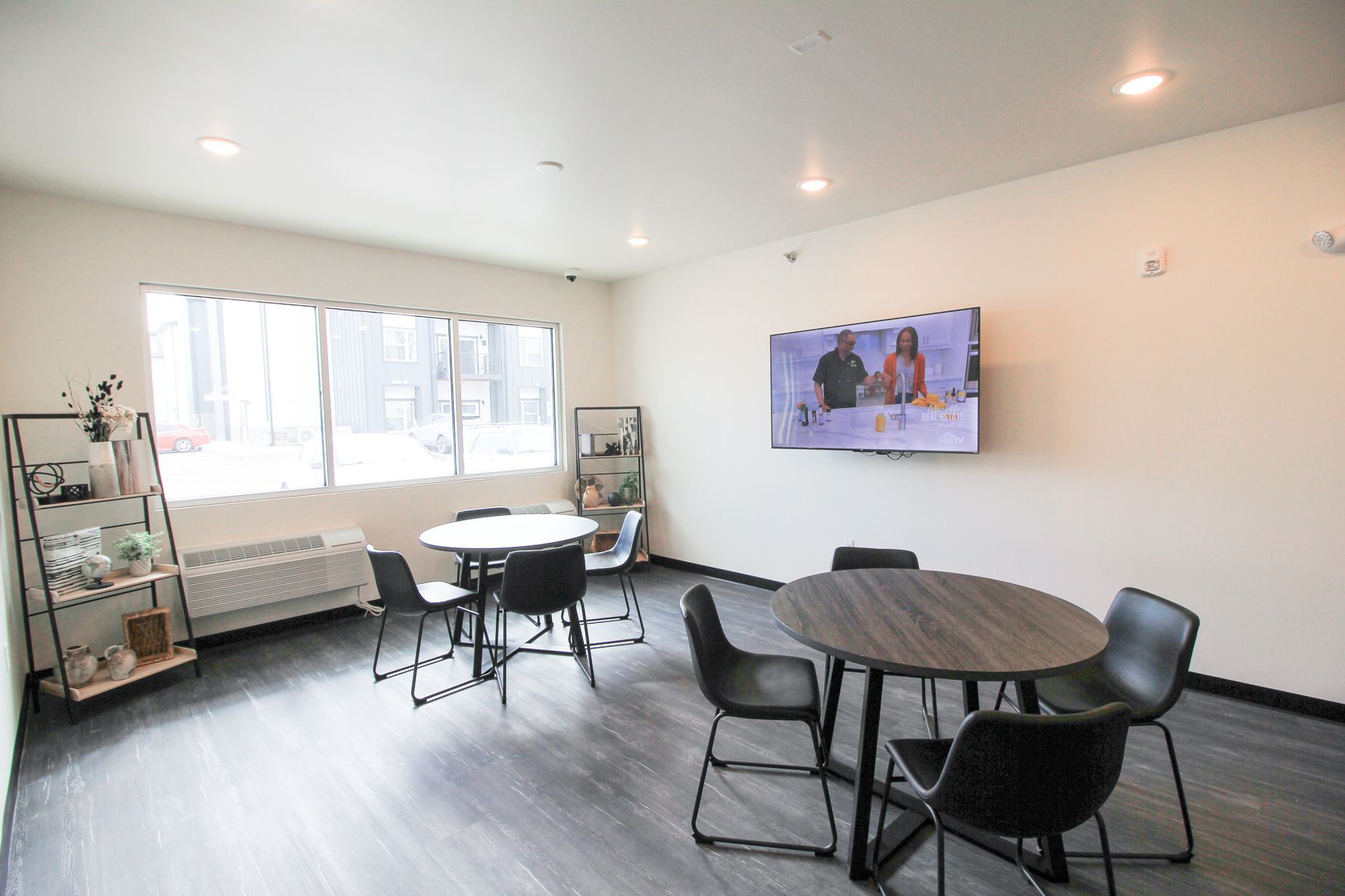 Bright communal lounge with round tables, chairs, a bench, wall TV, and large windows.